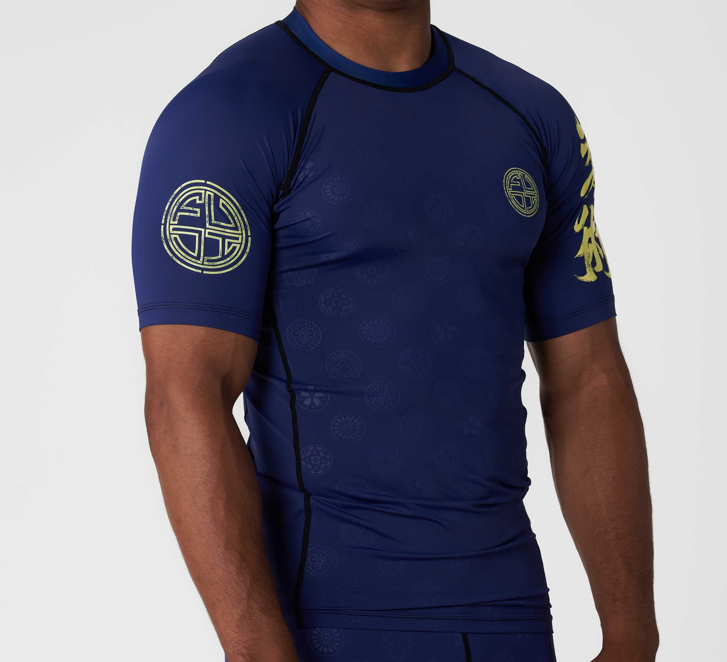 A person wears the Shogun Heat Gear Rashguard Navy, a fitted short-sleeve, navy athletic top featuring subtle circular patterns and gold emblem designs on the left sleeve and chest. The person's head is not visible.