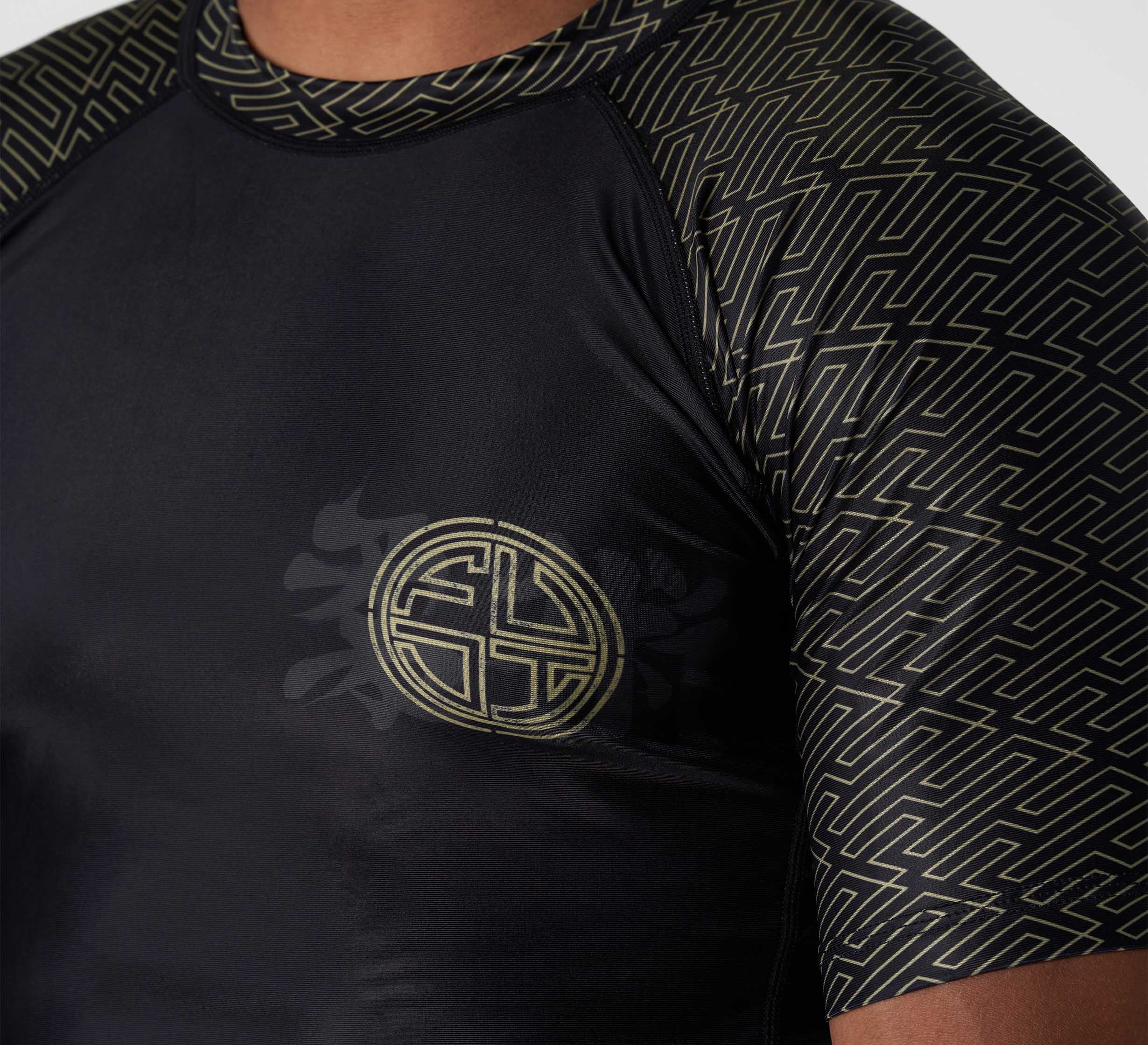 A close-up of a person wearing the Bushido Flex Lite Rashguard, featuring black fabric with gold geometric sleeve patterns and a circular gold maze emblem on the chest.