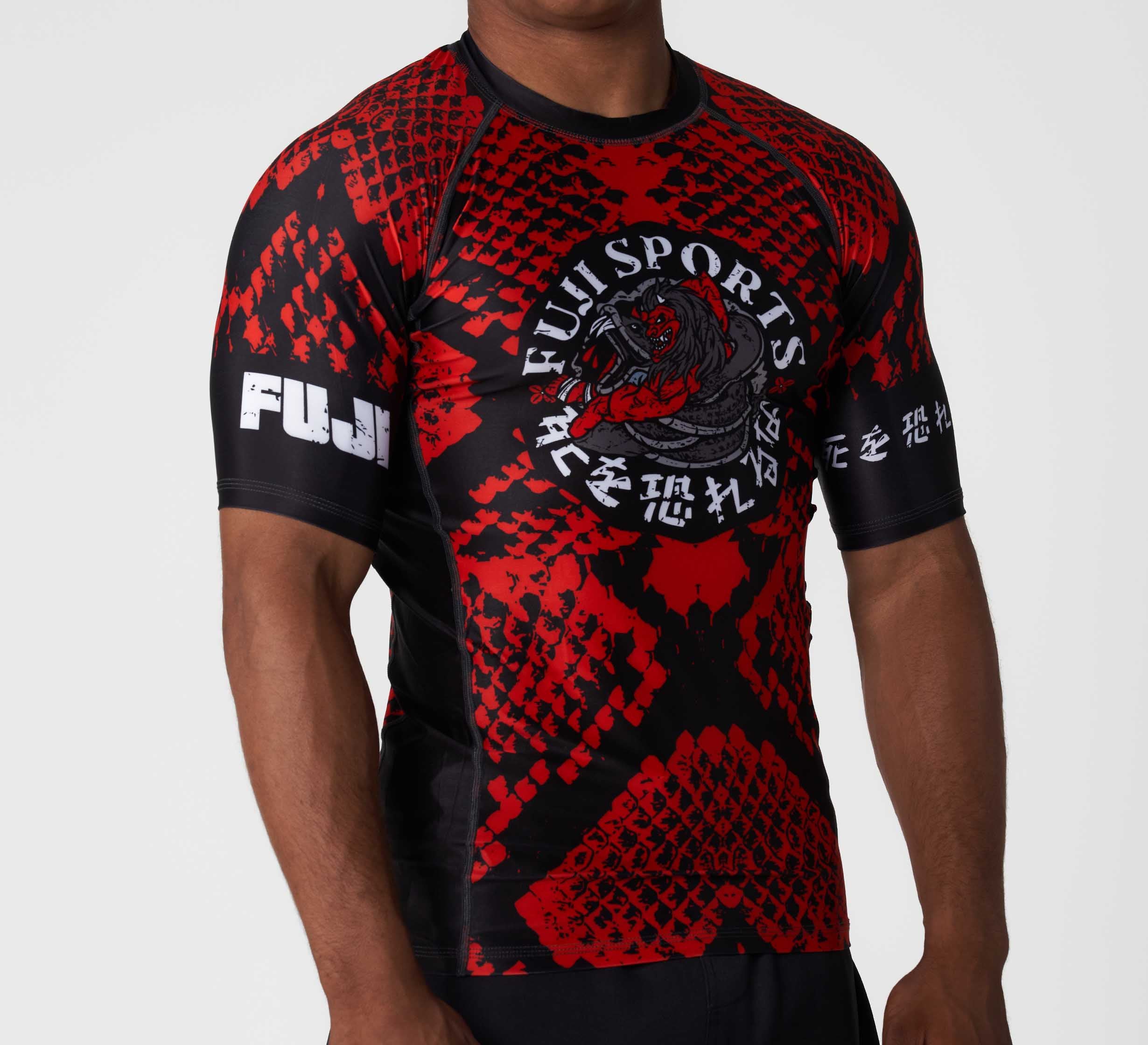 A person wears the Oni vs. Anaconda Short Sleeve Rashguard Red by Fuji Sports, featuring a red and black dragon design, white Japanese characters, and bold FUJI text on the sleeve, shown chest up against a plain background.