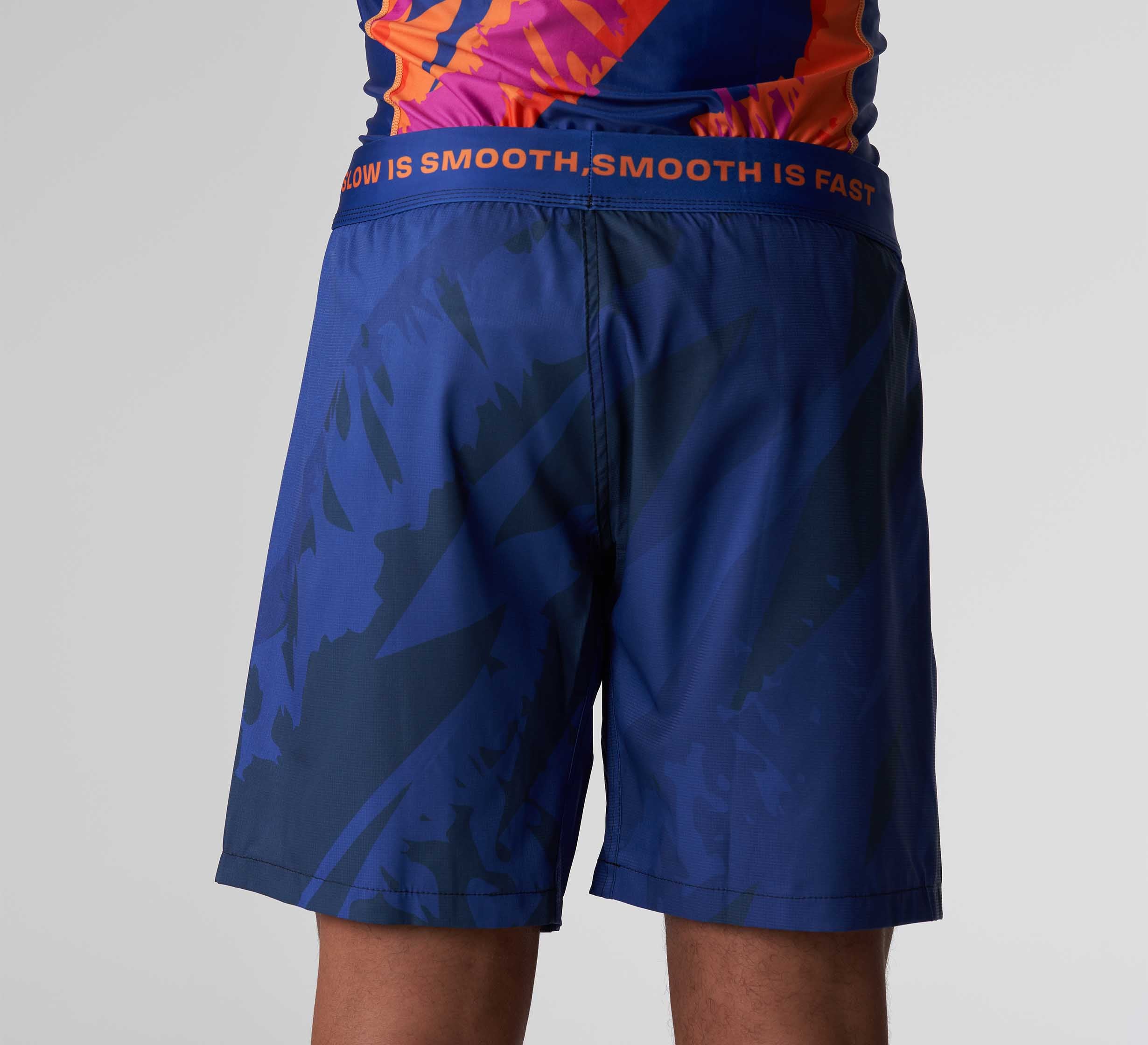 A person wearing Hanzo Flex Lite Shorts Blue with a patterned design and a colorful shirt, shown from the waist down and back. The waistband features "SMOOTH IS FAST, SLOW IS SMOOTH" in orange text.