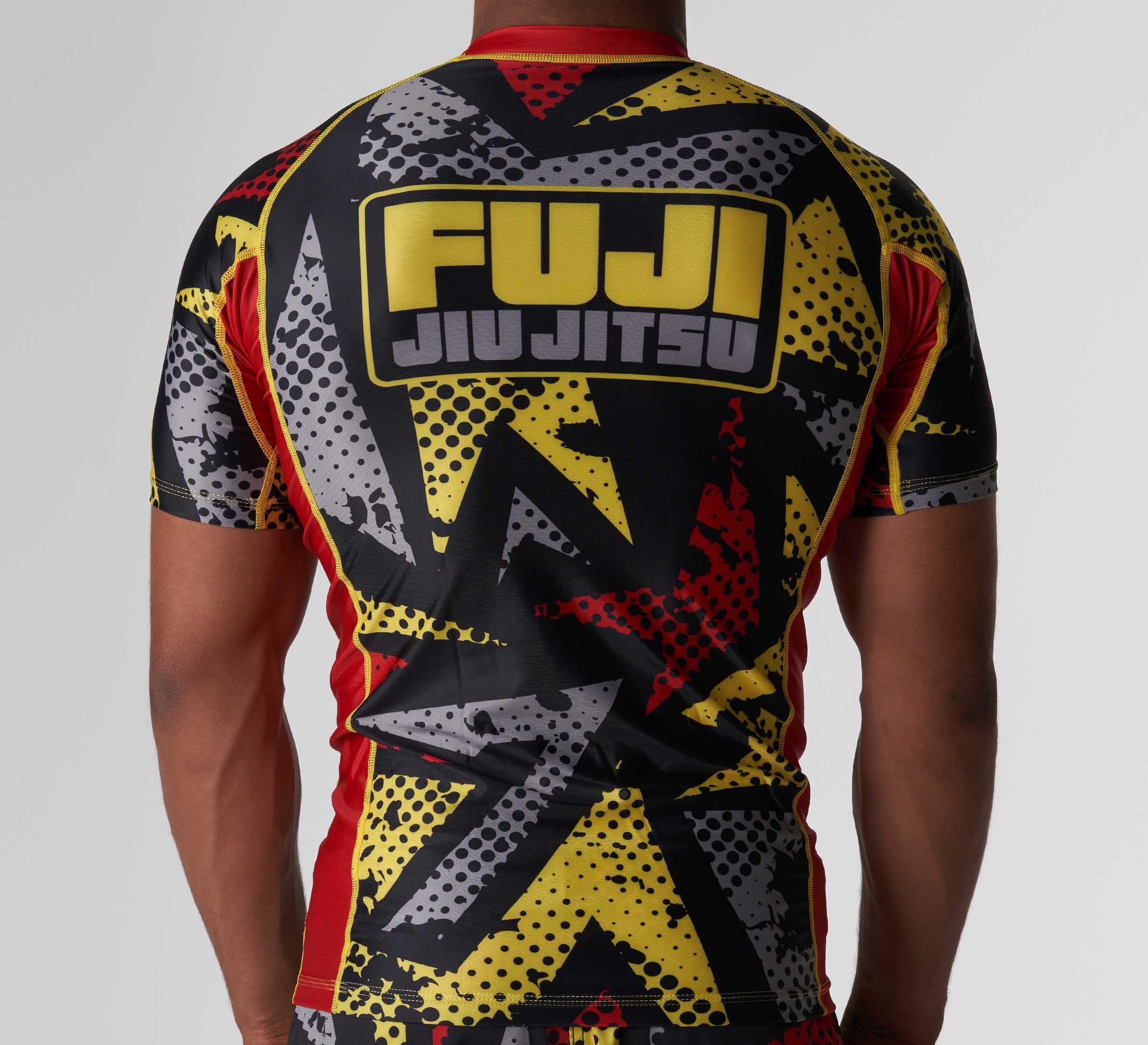 A person wearing the Shuriken Flex Lite Rashguard Black/Yellow with bold graphic patterns is seen from the back. The large FUJI JIU JITSU logo stands out on the upper back.