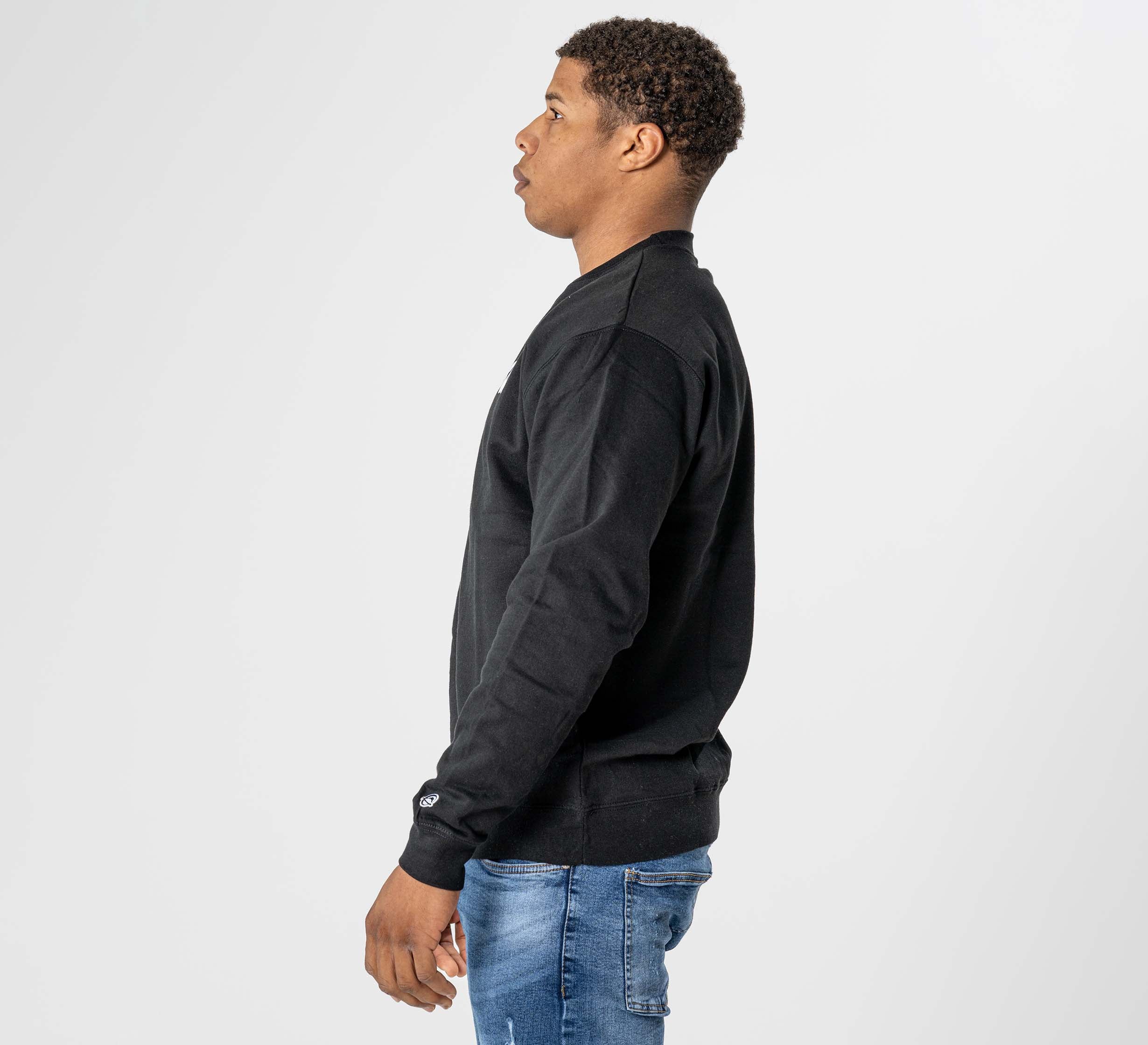 A man in profile, facing left, is wearing the Jiu Jitsu Player Crewneck Black with blue jeans against a plain white background.