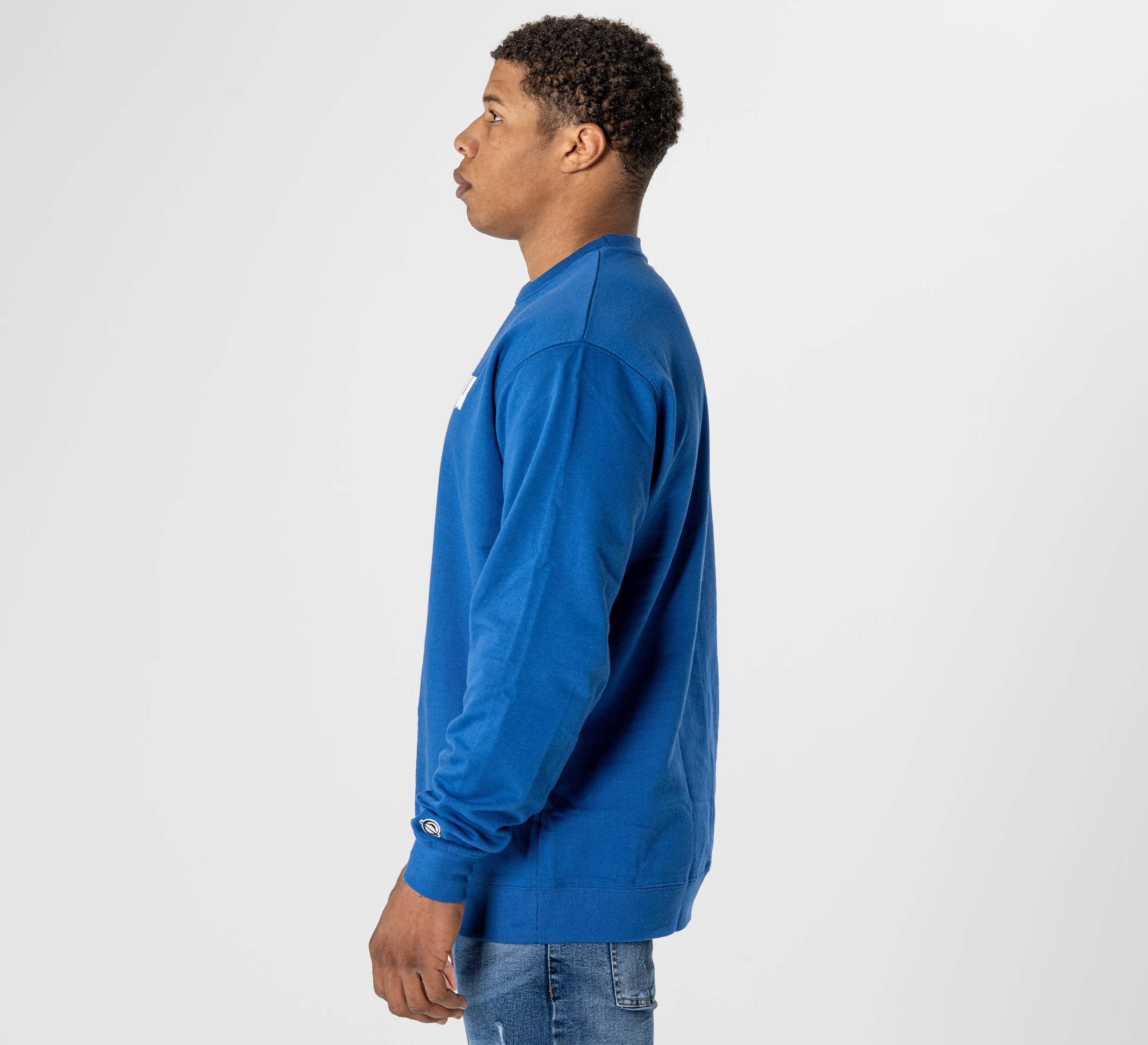 A man with short curly hair stands in profile facing left against a plain white background, wearing the Jiu Jitsu Player Crewneck Blue and light blue jeans.