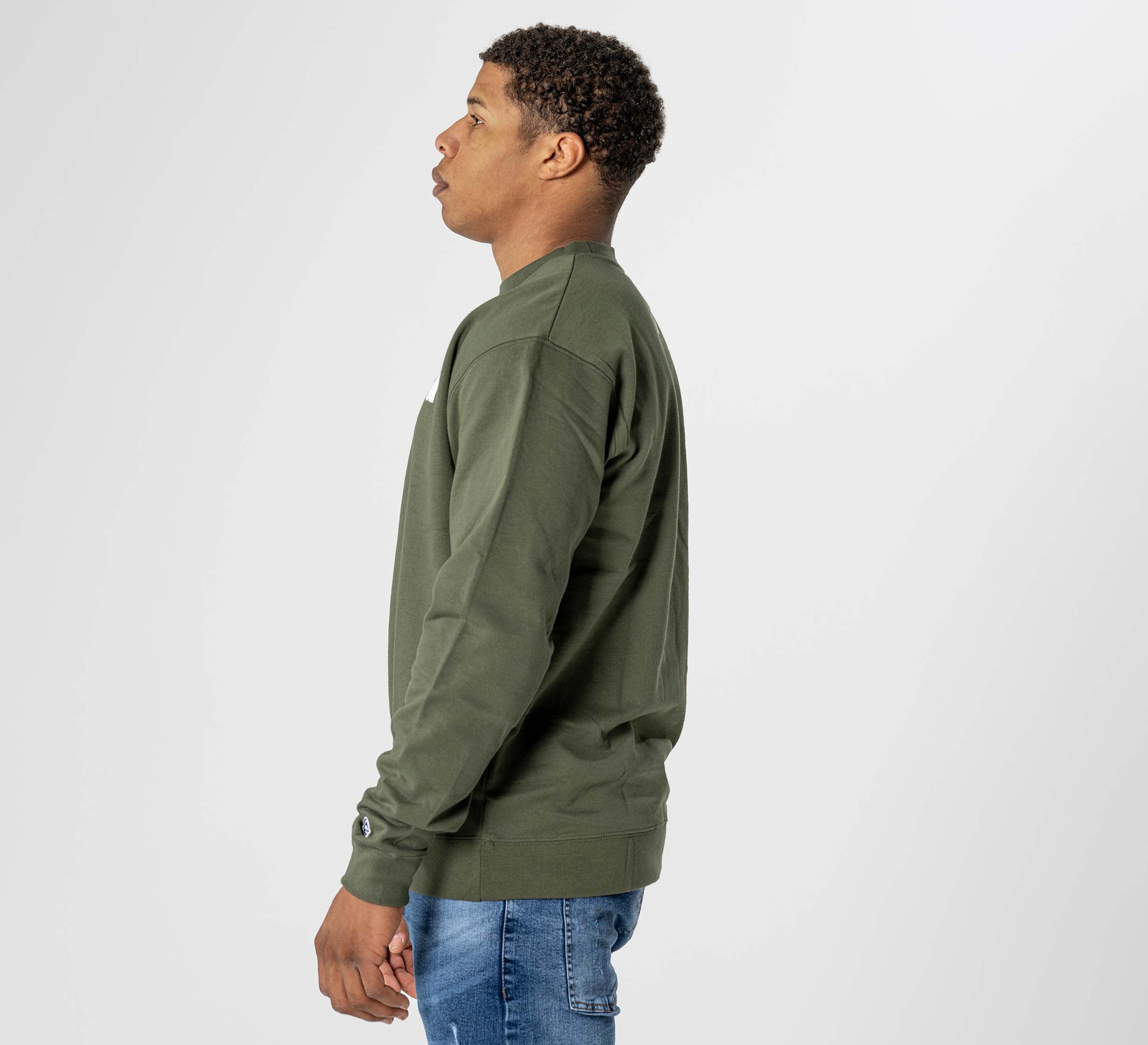 A man wears the Jiu Jitsu Player Crewneck Military Green with blue jeans, standing in profile against a white background, facing left and keeping his arms relaxed at his sides.