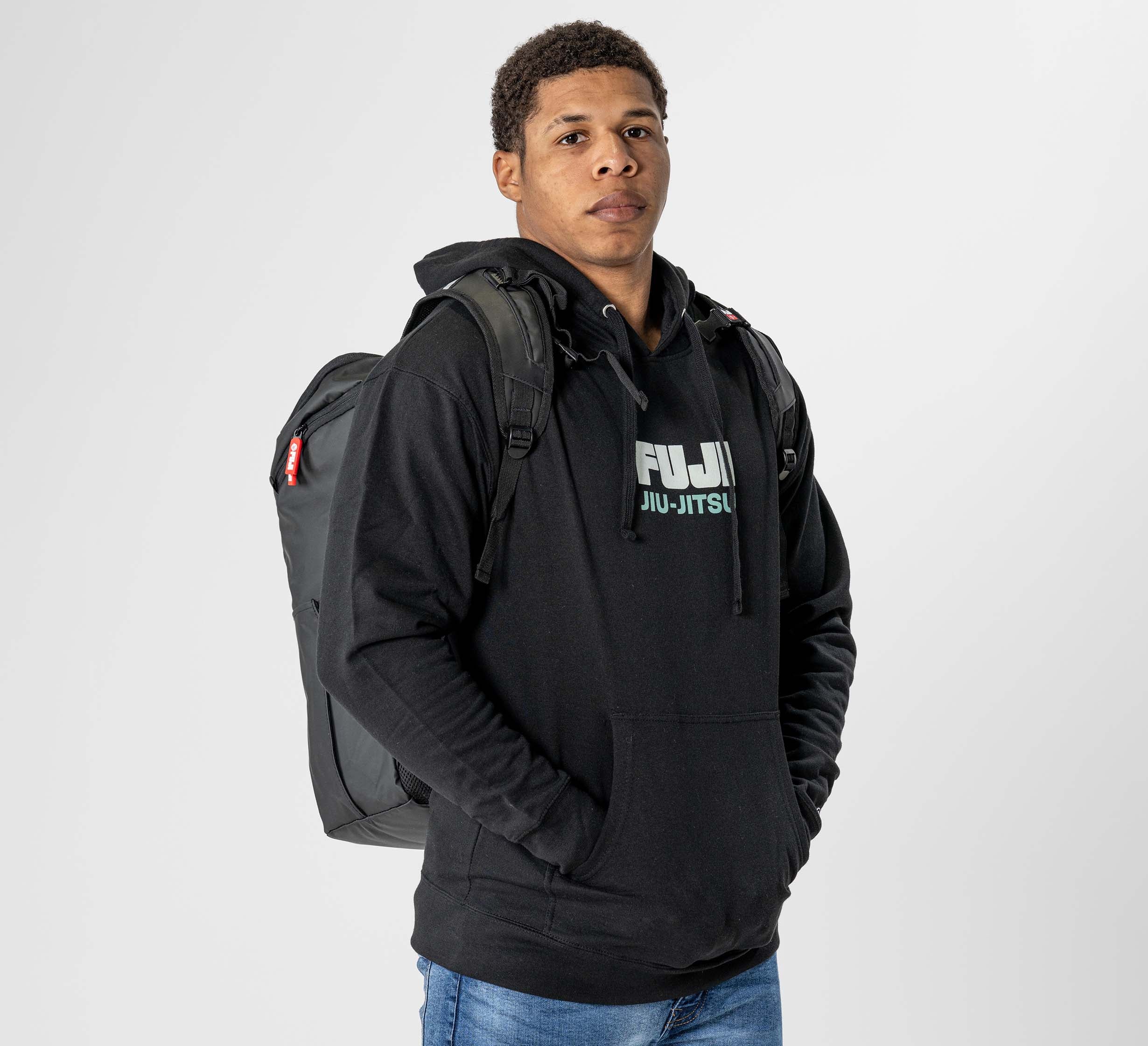 A young man stands facing forward against a plain white background, hands in pockets, wearing blue jeans, a black FUJI JIU-JITSU hoodie, and carrying the Urban Day Backpack Black.