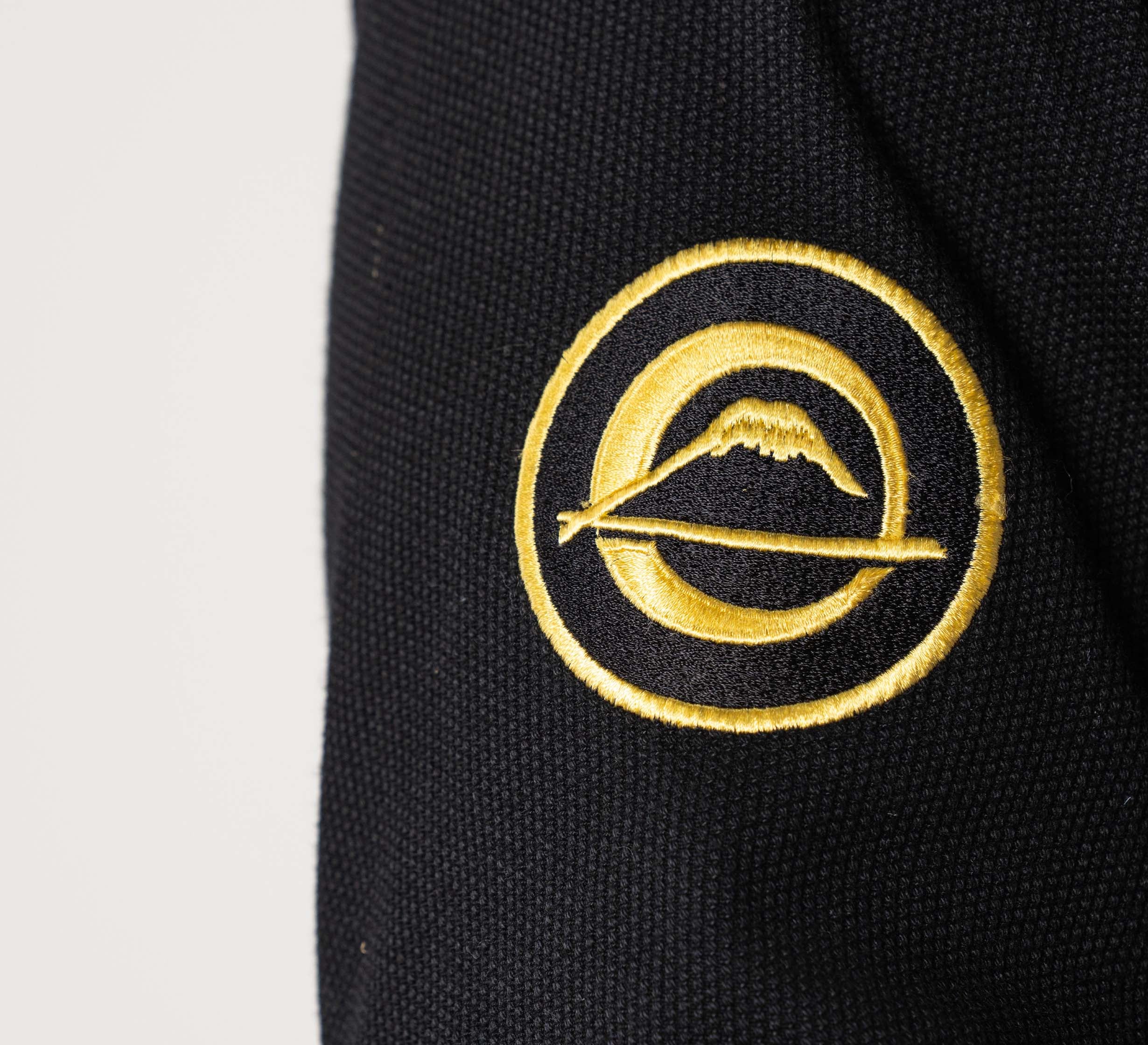 A close-up of the Flow-Tech BJJ Gi Black/Gold shows its black fabric with a yellow embroidered circular emblem featuring a mountain and a horizontal line.
