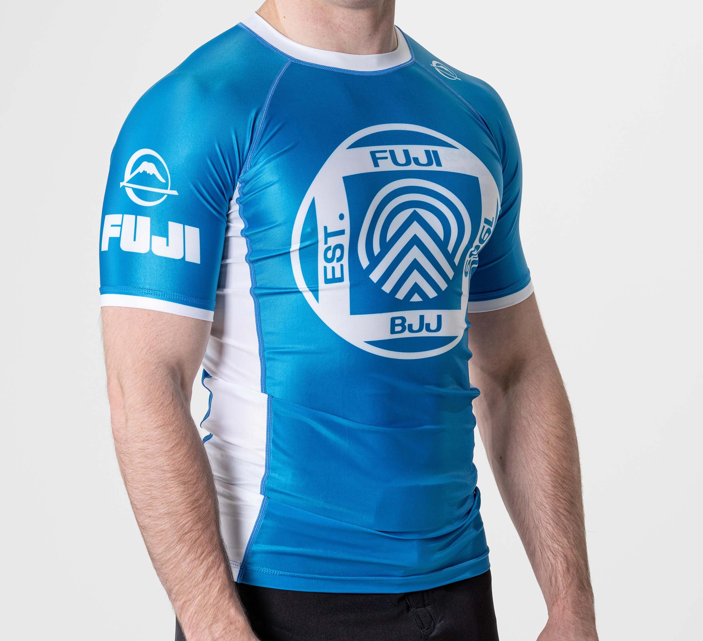 A person wearing the BJJ Flex Lite Rashguard Blue, featuring logos and text on the chest, sleeve, and side, stands against a plain white background.