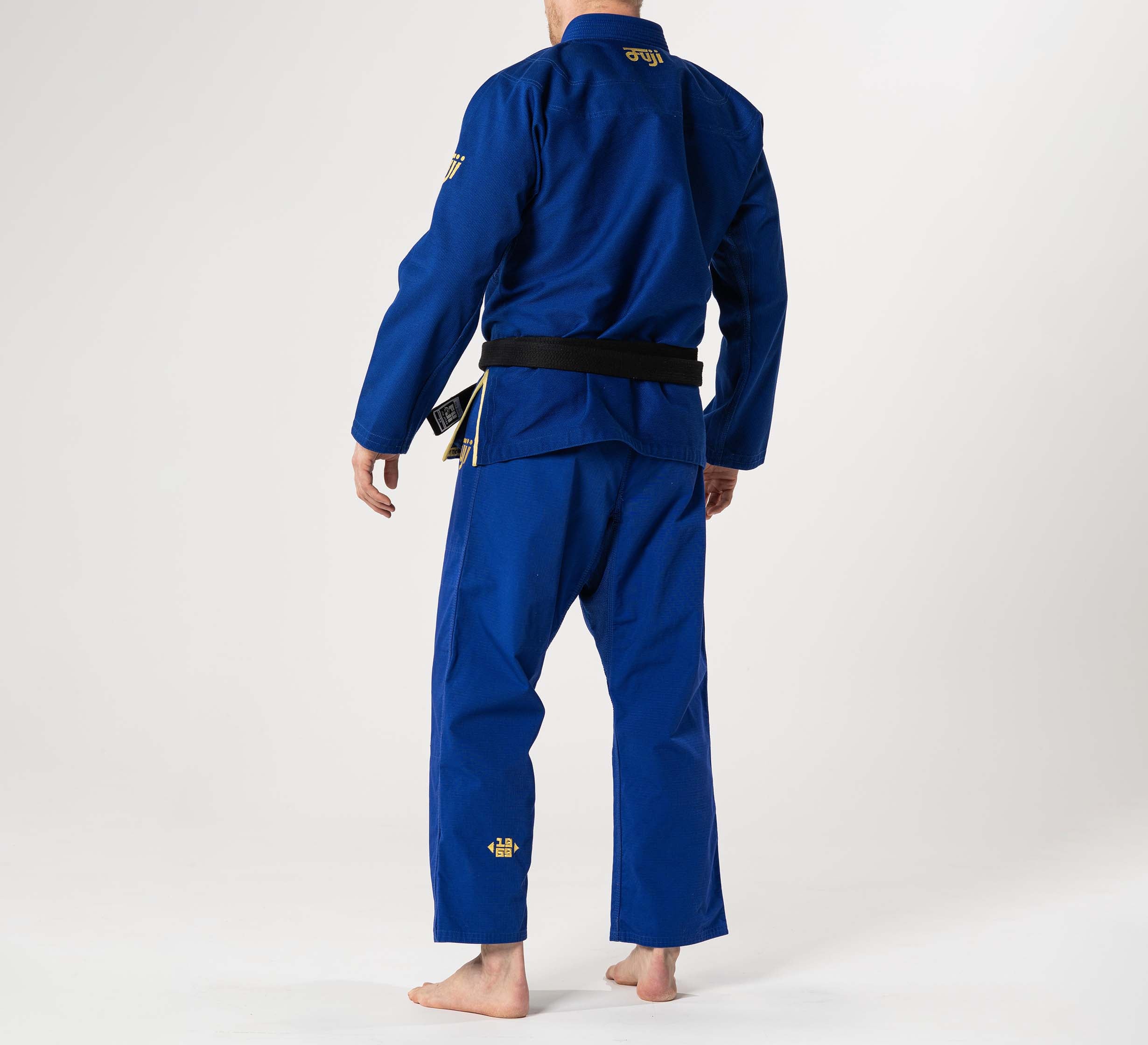 A person stands barefoot on a white background, wearing the Flow-Tech BJJ Gi Blue/Gold with a black belt. The individual faces away from the camera, displaying the back of the uniform.