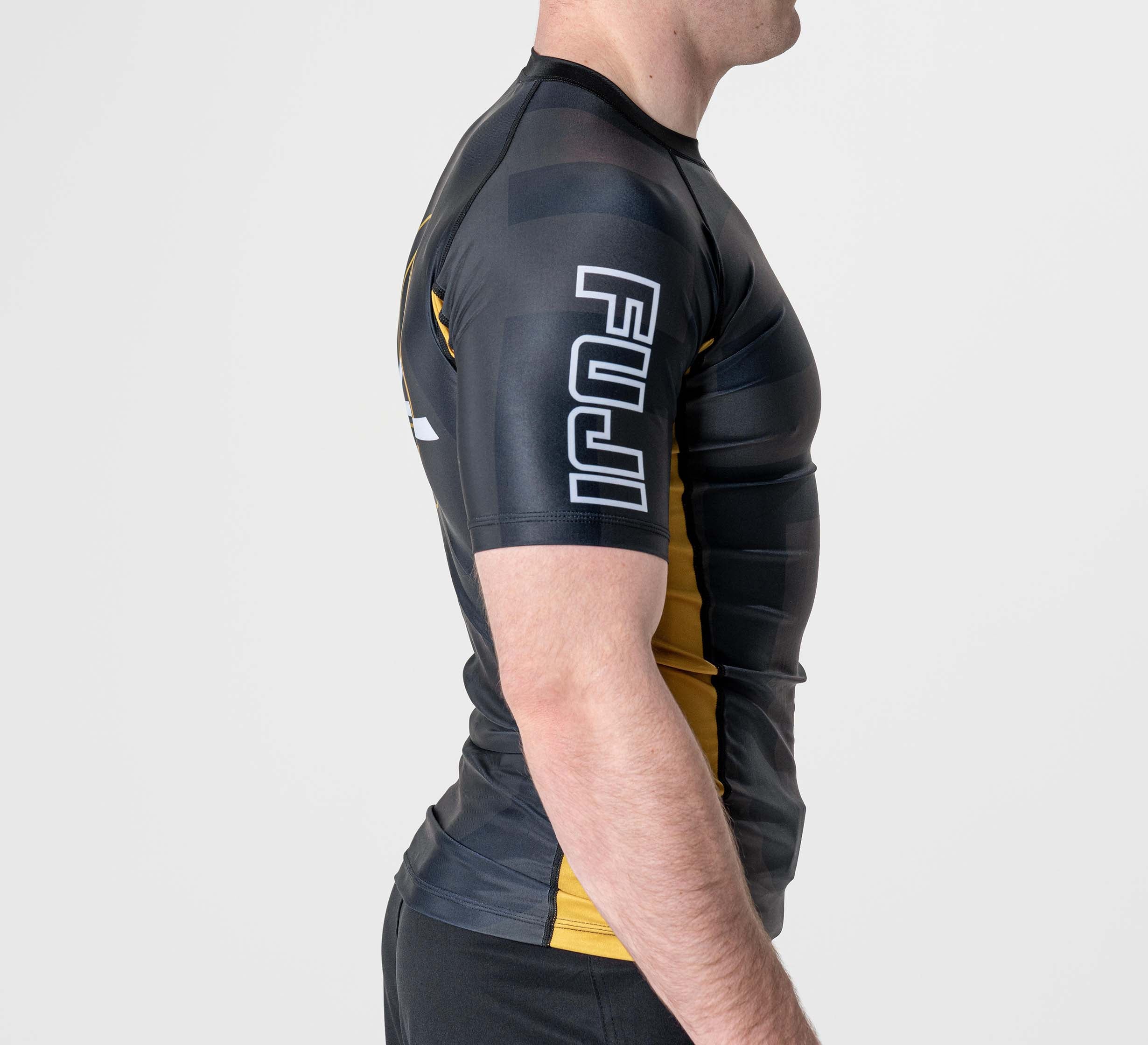 A person wearing the Rebel Flex Lite Rashguard Black with short sleeves and yellow accents, featuring "FUJI" in white on the upper sleeve, shown from the side against a plain white background.