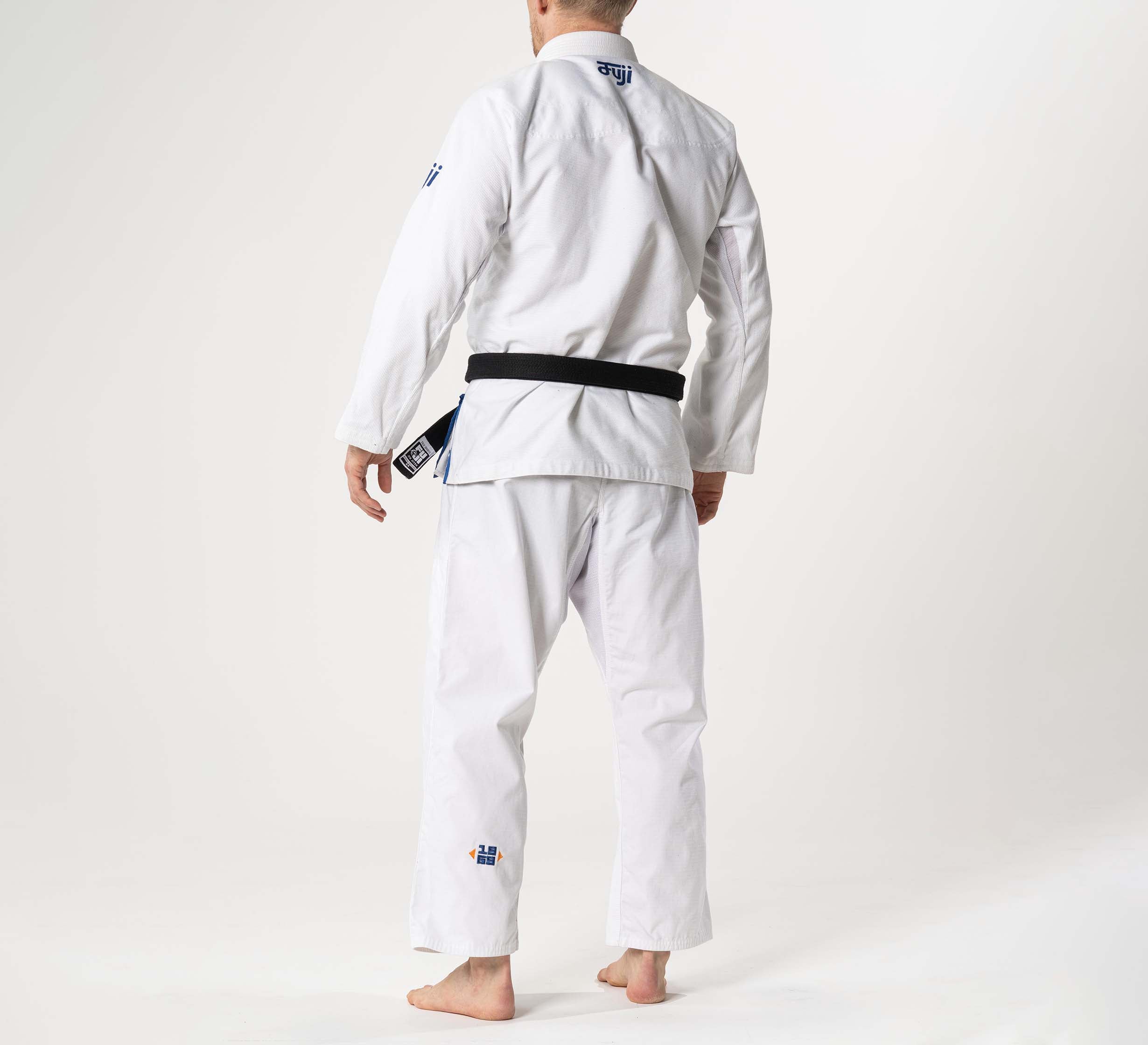 A person wears the Flow-Tech BJJ Gi in white, navy, and orange with a black belt, standing barefoot on a plain light background, facing away from the camera.