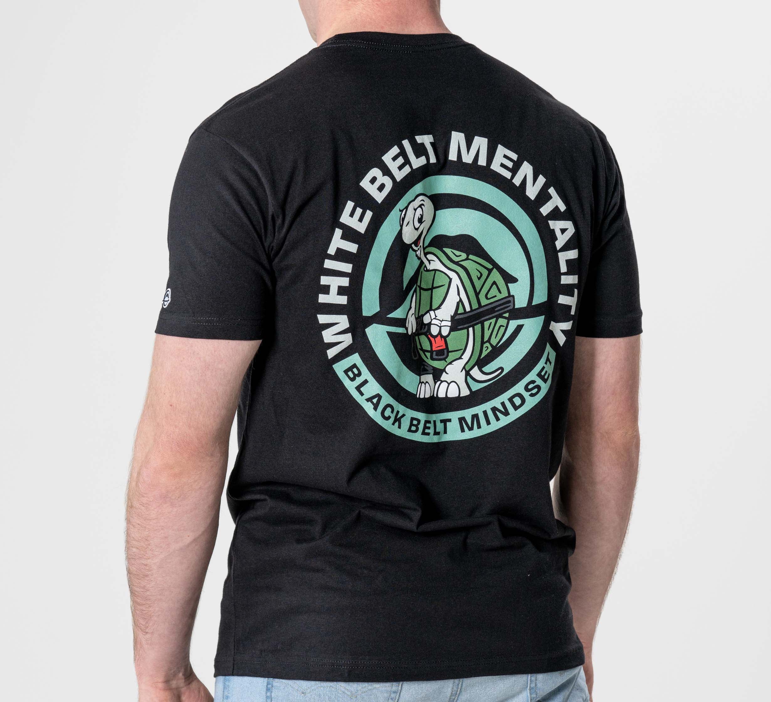 Someone wearing the Black Belt Mentality T-Shirt Black, featuring a graphic of a turtle holding a black belt and the phrase "White Belt Mentality, Black Belt Mindset" in a circular design on the back.
