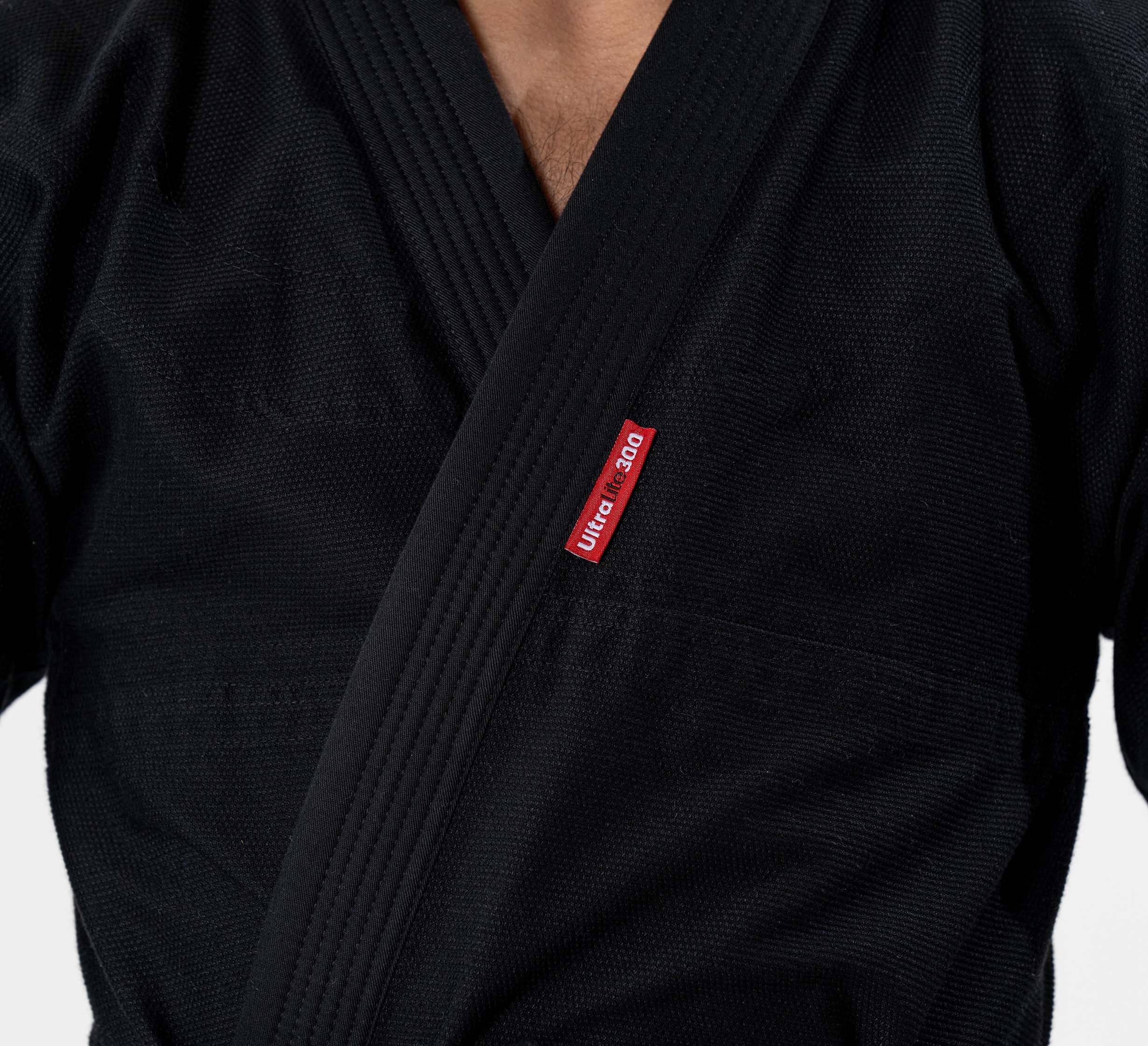 A child wears the Kids Ultra Lite 300 BJJ Gi in black, featuring a visible red front tag; the photo is cropped to highlight the upper torso and part of the neck.