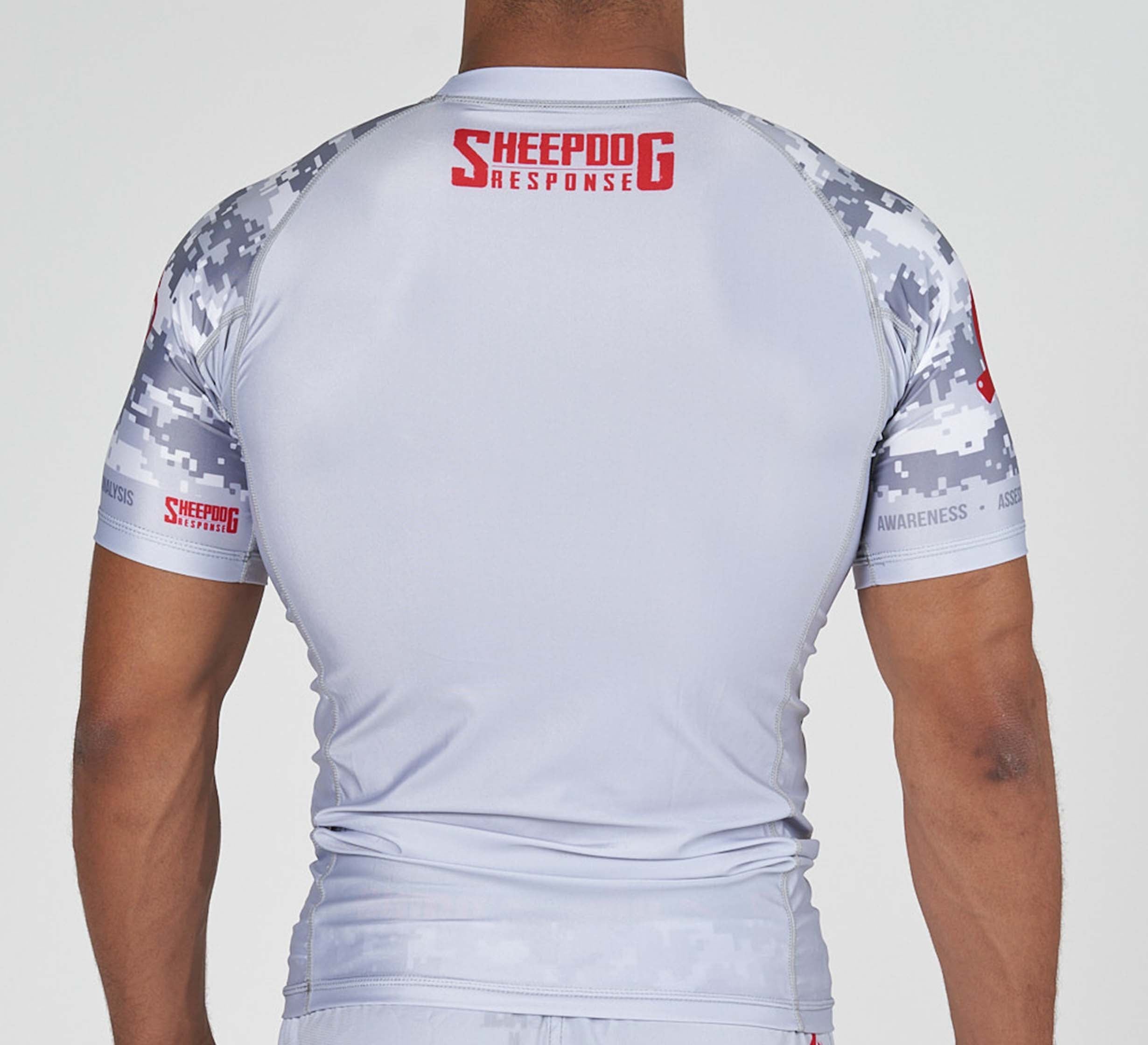 A person is seen from the back wearing the Sheepdog Response Flex Lite Rashguard Grey, a fitted athletic shirt featuring a gray camouflage design and red SHEEPDOG RESPONSE text on the upper back and sleeves, highlighting their muscular build.