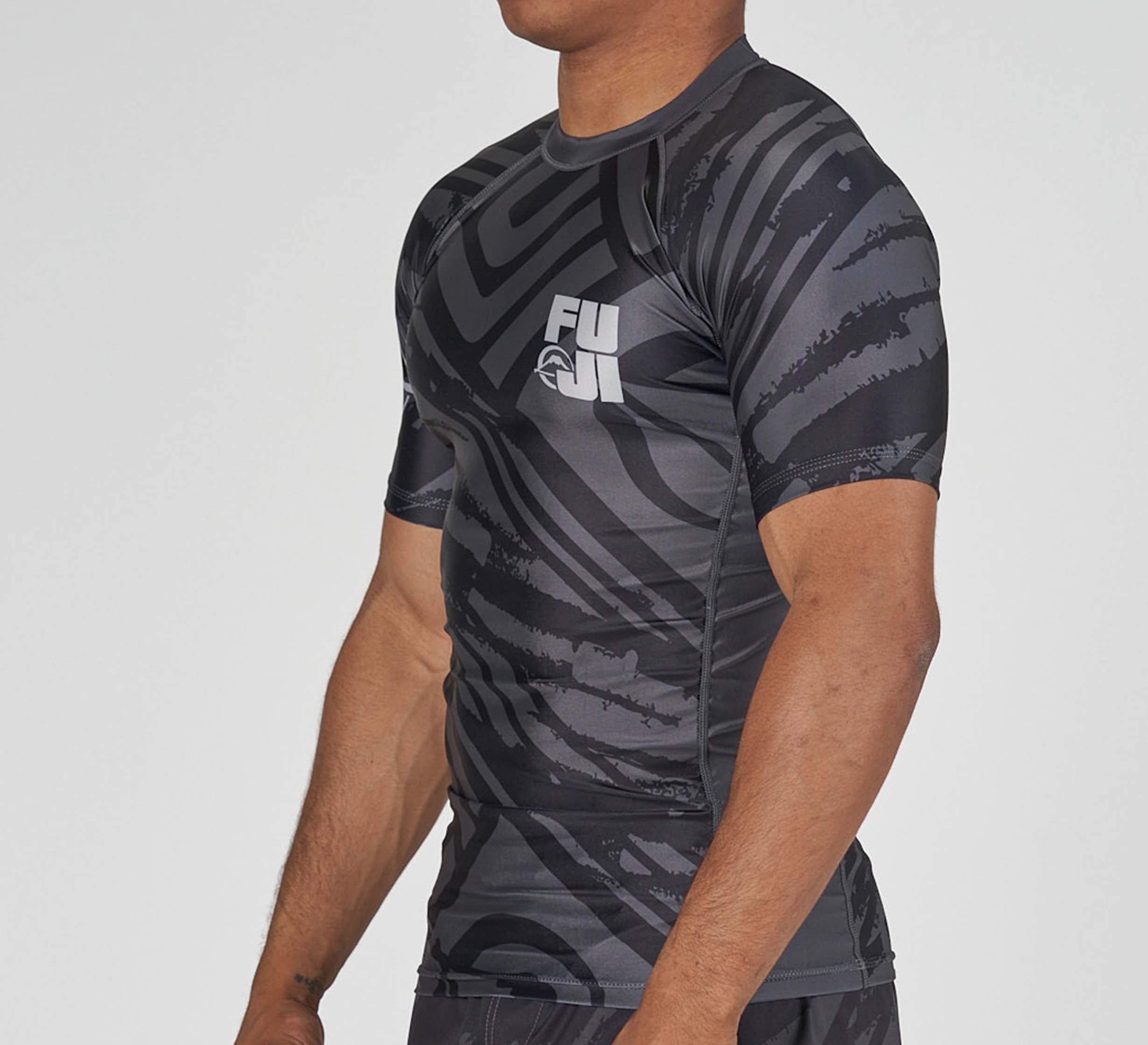 A person wearing the Battle Flex Lite Rashguard Black, a short-sleeved athletic top with a black and gray pattern and "FUJI" on the chest, stands against a plain light background.