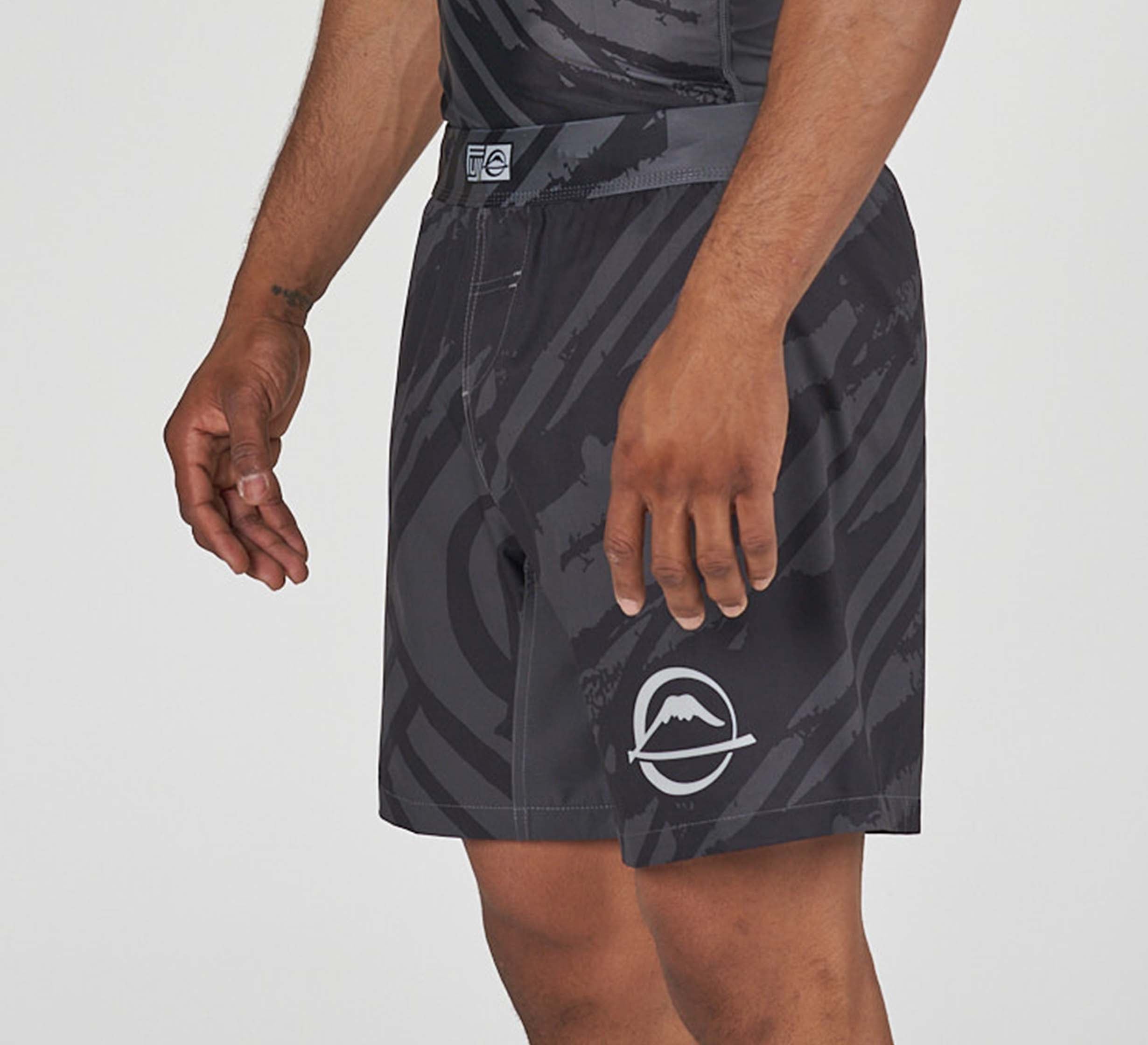 A person wears Battle Flex Lite Shorts Black, featuring a black and gray pattern and a white mountain logo on the left leg, standing against a plain background.