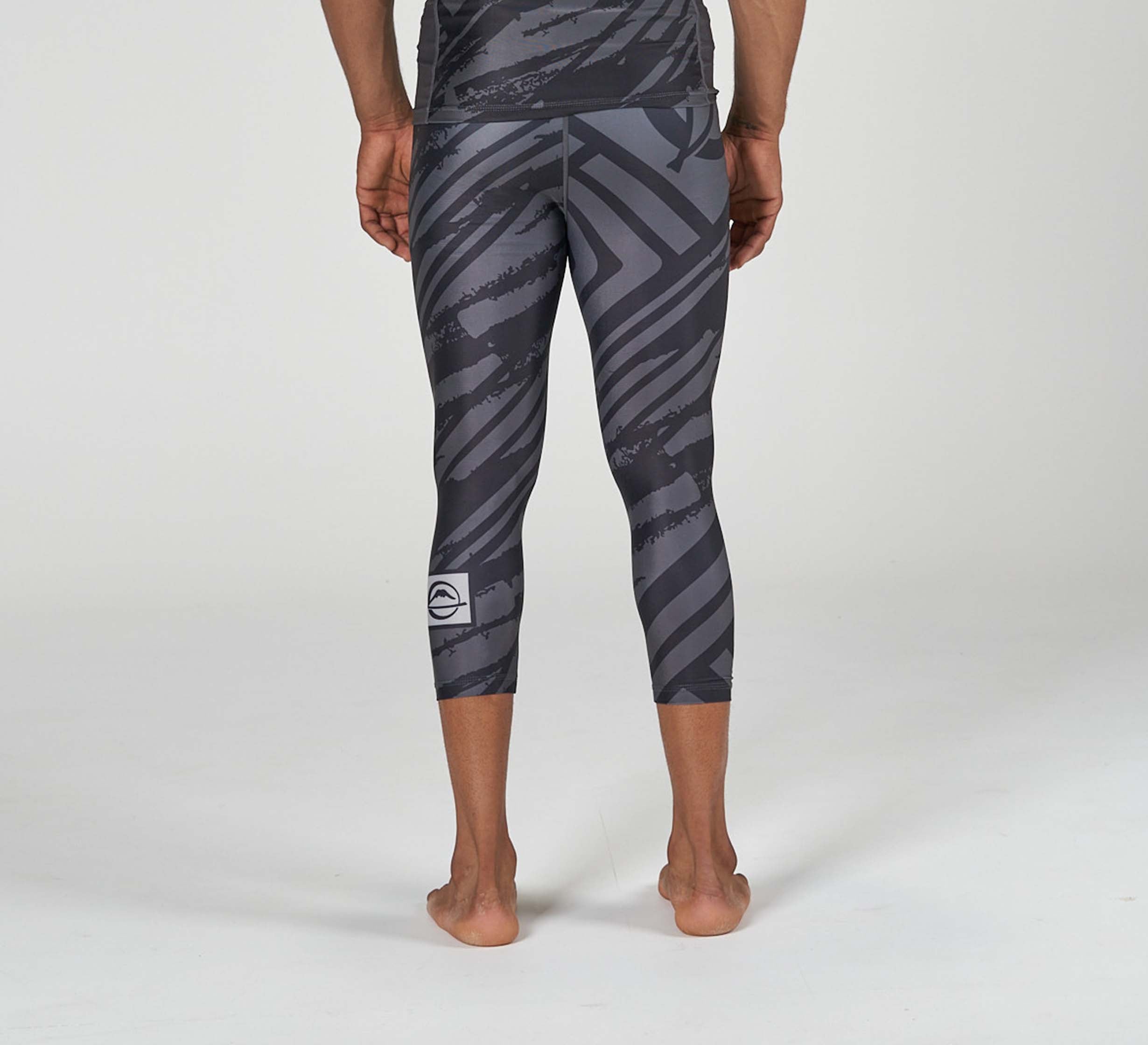 A person stands barefoot facing away, wearing Battle Flex Lite Spat Black leggings with bold diagonal stripes and a small white logo on the left calf. The mid-calf leggings feature a dark gray pattern against a plain, light background.