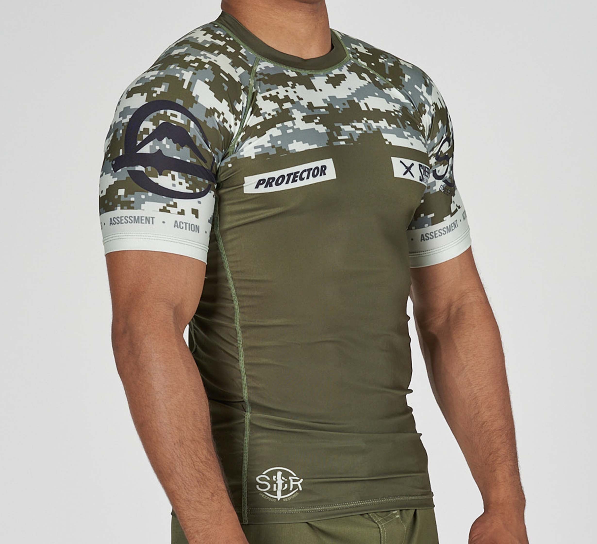 A person wears the Sheepdog Response Flex Lite Rashguard in Military Green, featuring olive and camo patterns with "PROTECTOR," "ASSESSMENT," "ACTION," and several logos, standing against a plain background.