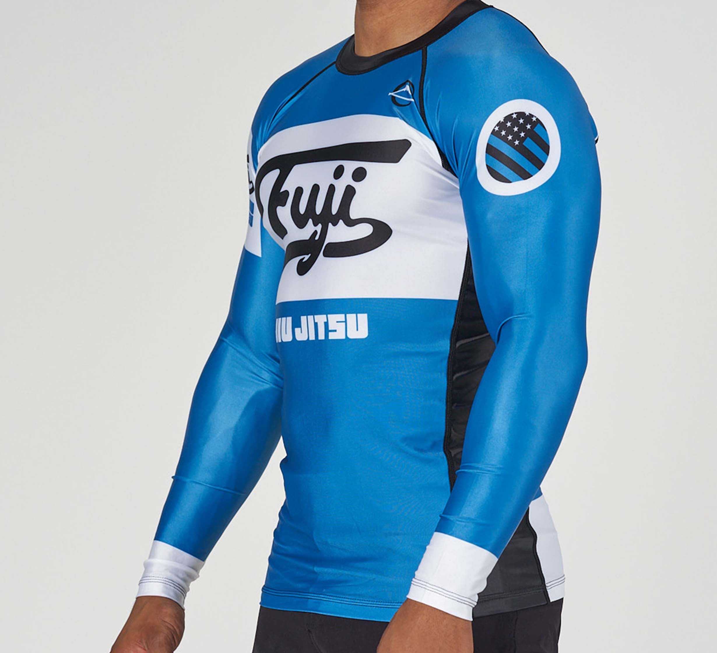 A person wearing the Jiu Jitsu Script Flex Lite Long Sleeve Rashguard Blue, featuring blue, black, and white colors, "Kuji Jiu Jitsu" text, and a circular American flag patch on the upper arm. The person's head is not visible.