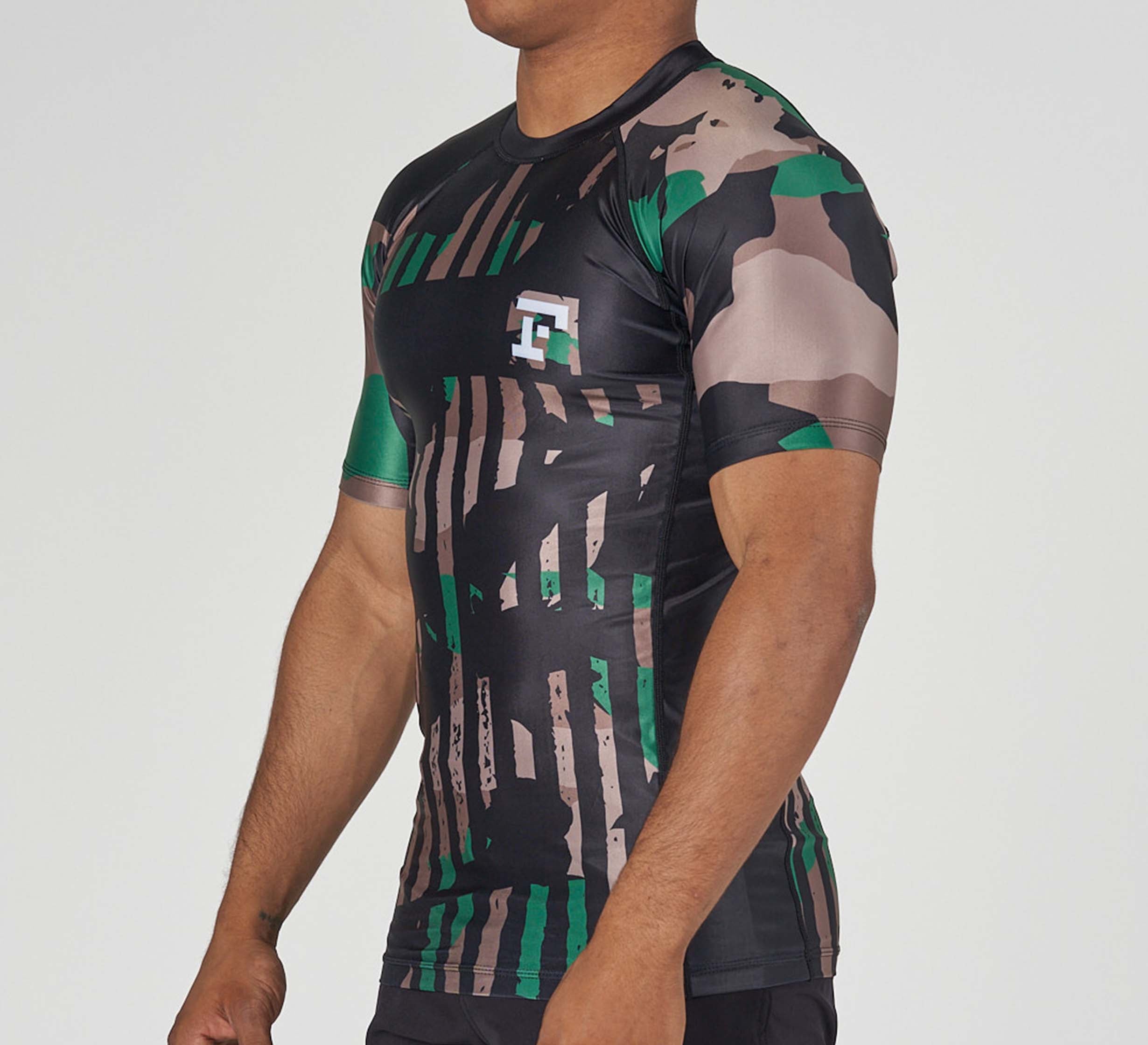 Someone wearing the Camo Flex Lite Rashguard, a fitted short-sleeved athletic shirt with a green, black, brown, and beige camo pattern and a small white logo on the chest, set against a plain light background.