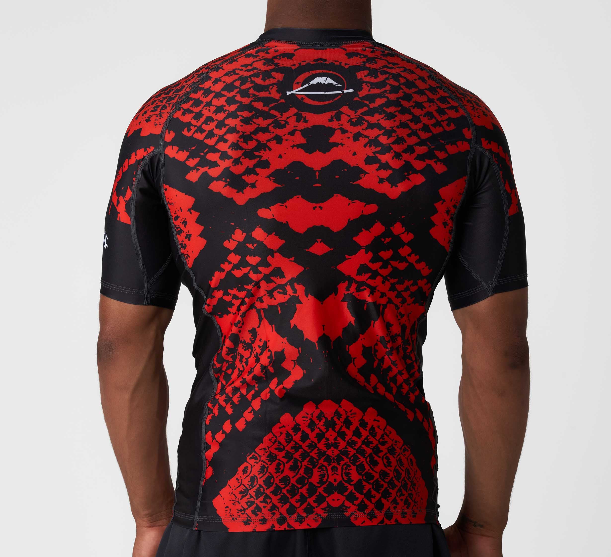 A person faces away from the camera, wearing the Oni vs. Anaconda Short Sleeve Rashguard Red—an athletic black and red shirt with a bold, symmetrical, scale-like pattern. The short-sleeve rashguard stands out against a plain white background.