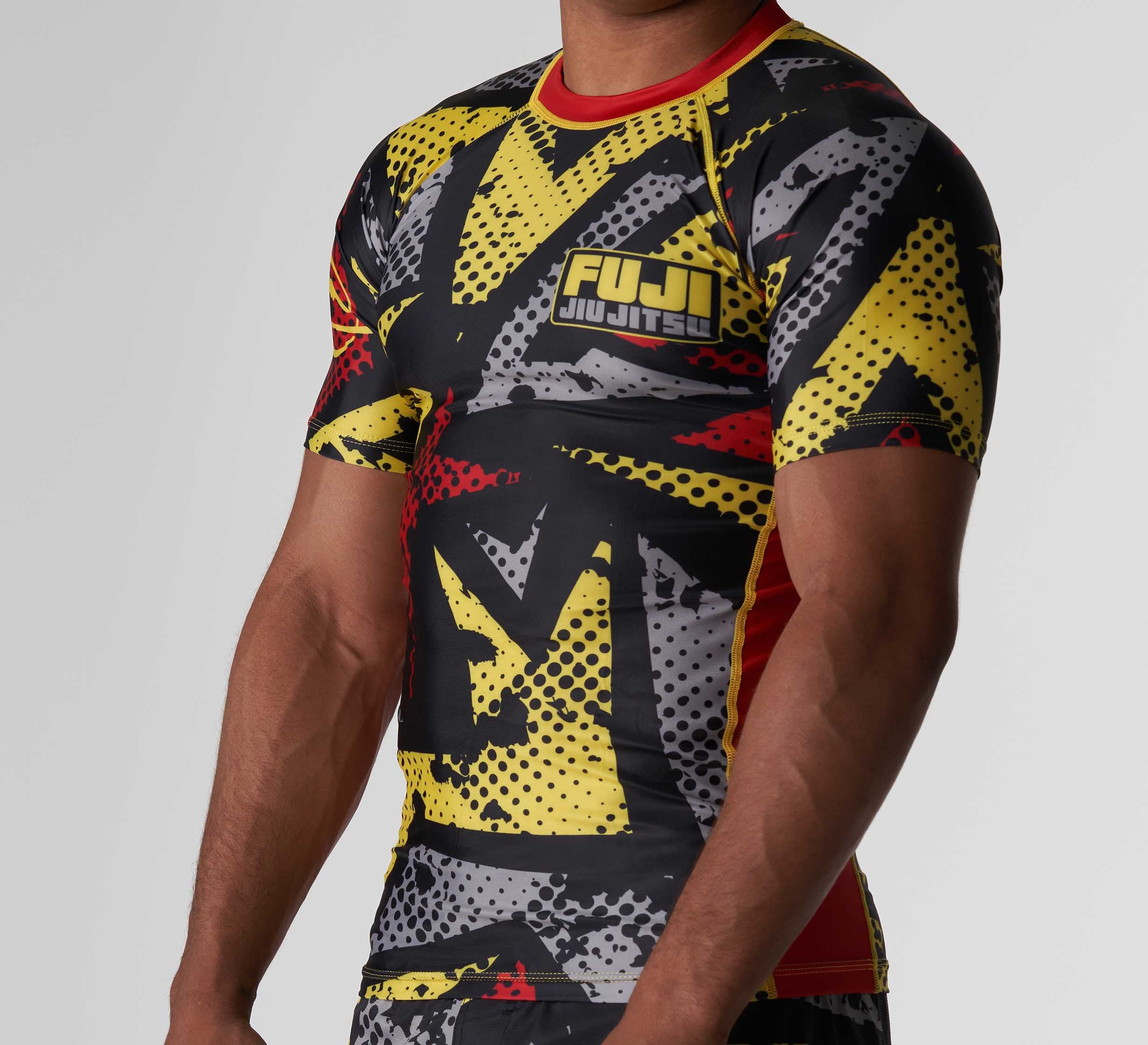 A person is wearing the Shuriken Flex Lite Rashguard Black/Yellow, a short-sleeved, form-fitting top with a bold abstract pattern in yellow, black, red, and white and "FUJI JIU JITSU" printed on the chest. The face is not visible.