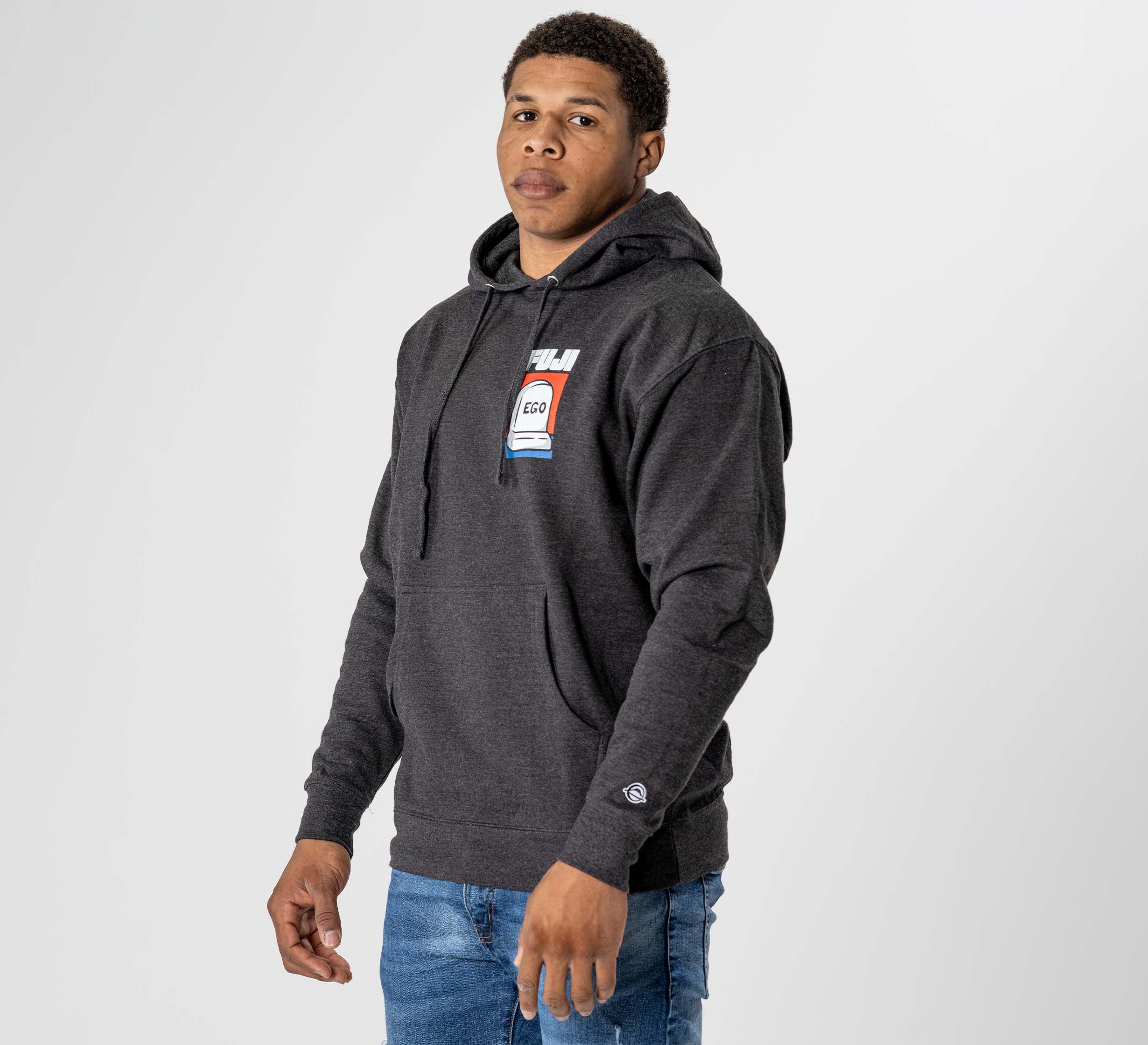 A man wearing the Submit Your Ego Hoodie in heather black, featuring a chest graphic, stands facing forward against a plain light background with one arm slightly bent. He pairs the hoodie with blue jeans.