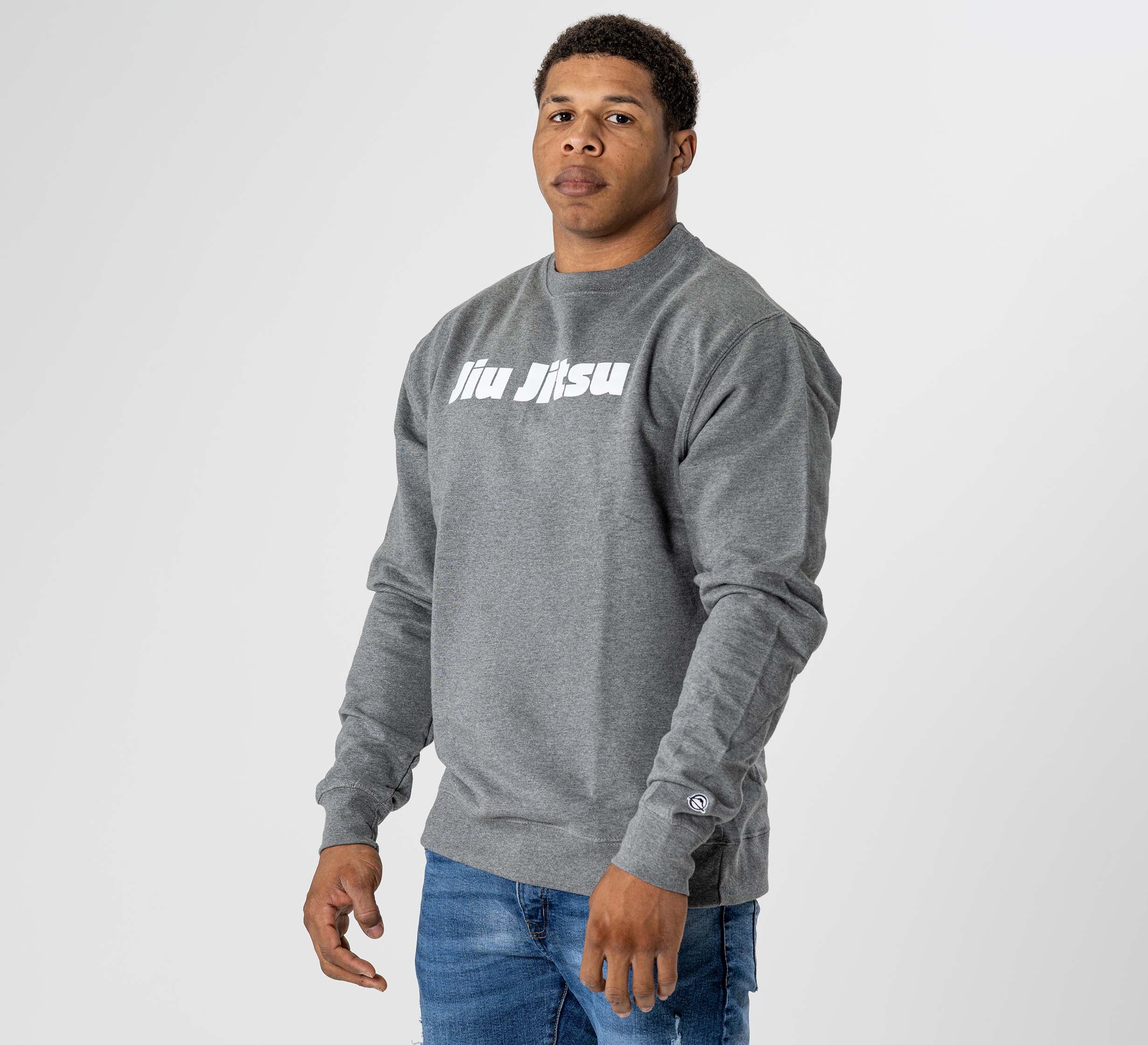 A man in the Jiu Jitsu Player Crewneck Heather Grey, featuring white "jiu jitsu" text on the chest, stands against a plain light gray background. He looks slightly to the side with a neutral expression and wears blue jeans.