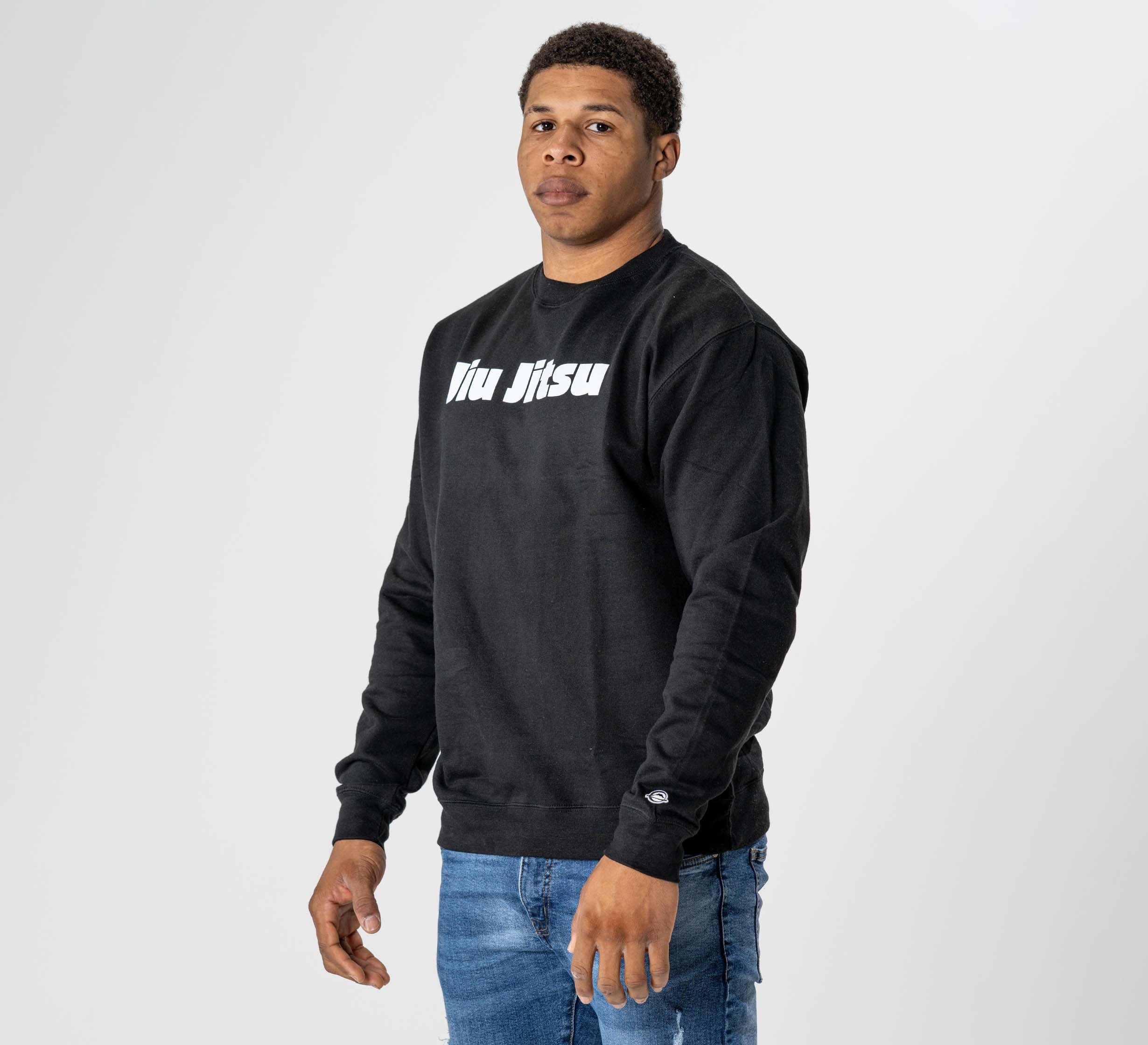 A man wears the Jiu Jitsu Player Crewneck Black, featuring "Jiu Jitsu" text, paired with blue jeans. He stands against a plain white background, looking slightly to the side.