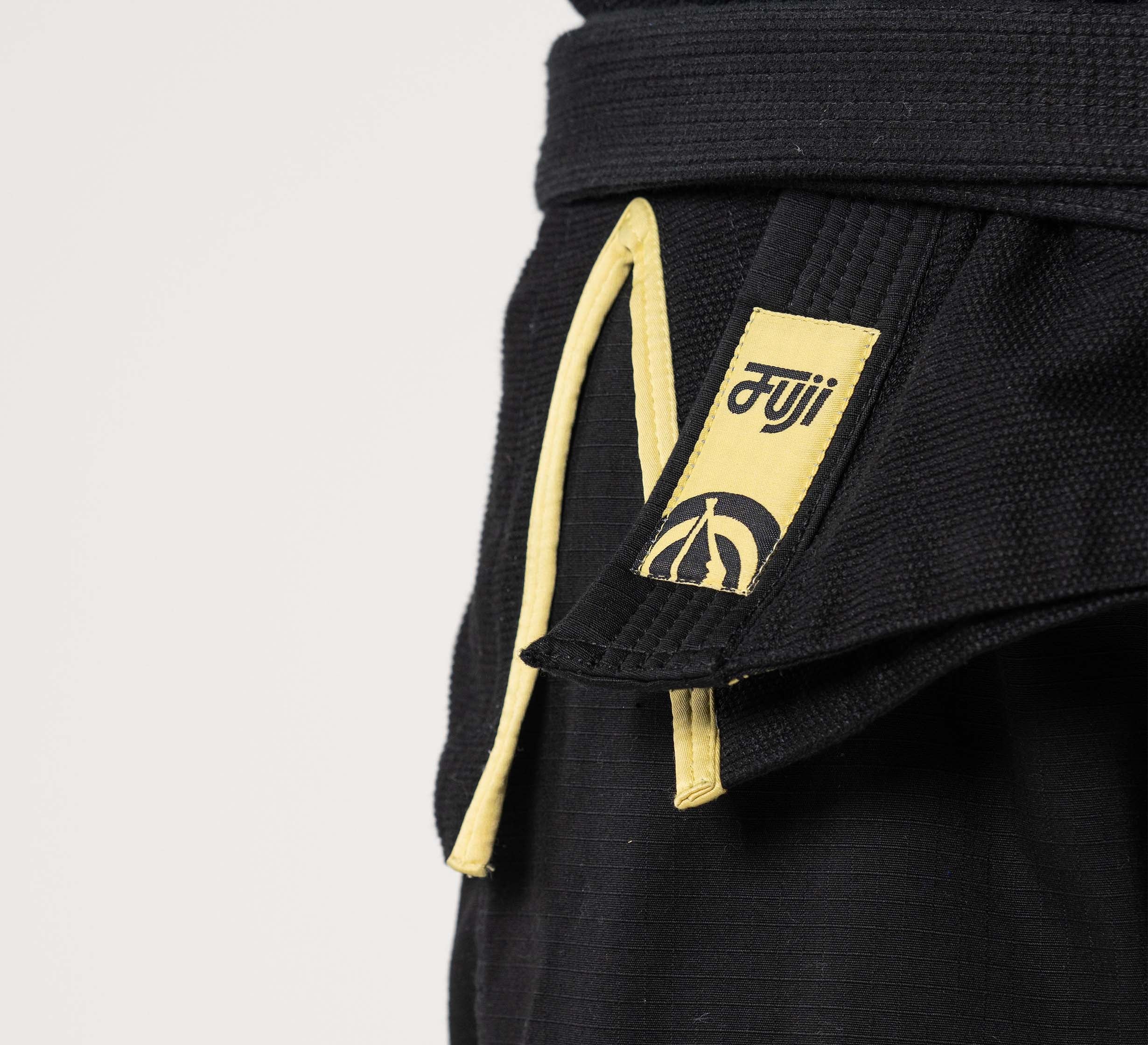 Close-up of a person wearing the Flow-Tech BJJ Gi Black/Gold, featuring a yellow-edged belt, Fuji brand tag, and part of a circular logo. The background is plain and light-colored.