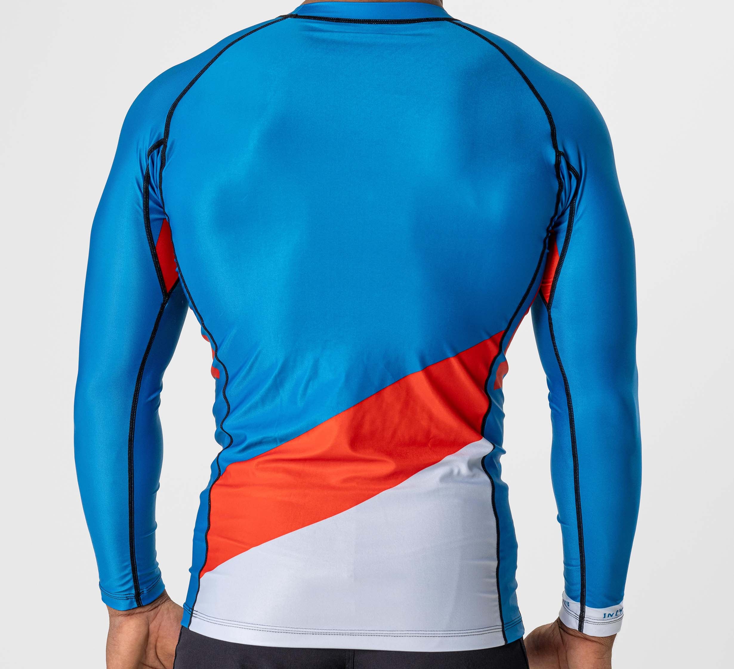 A person is seen from behind wearing the Pacer Flex Lite Long Sleeve Rashguard Blue/Red, a form-fitting athletic shirt with black seams and blue fabric featuring diagonal red and white blocks near the lower back.