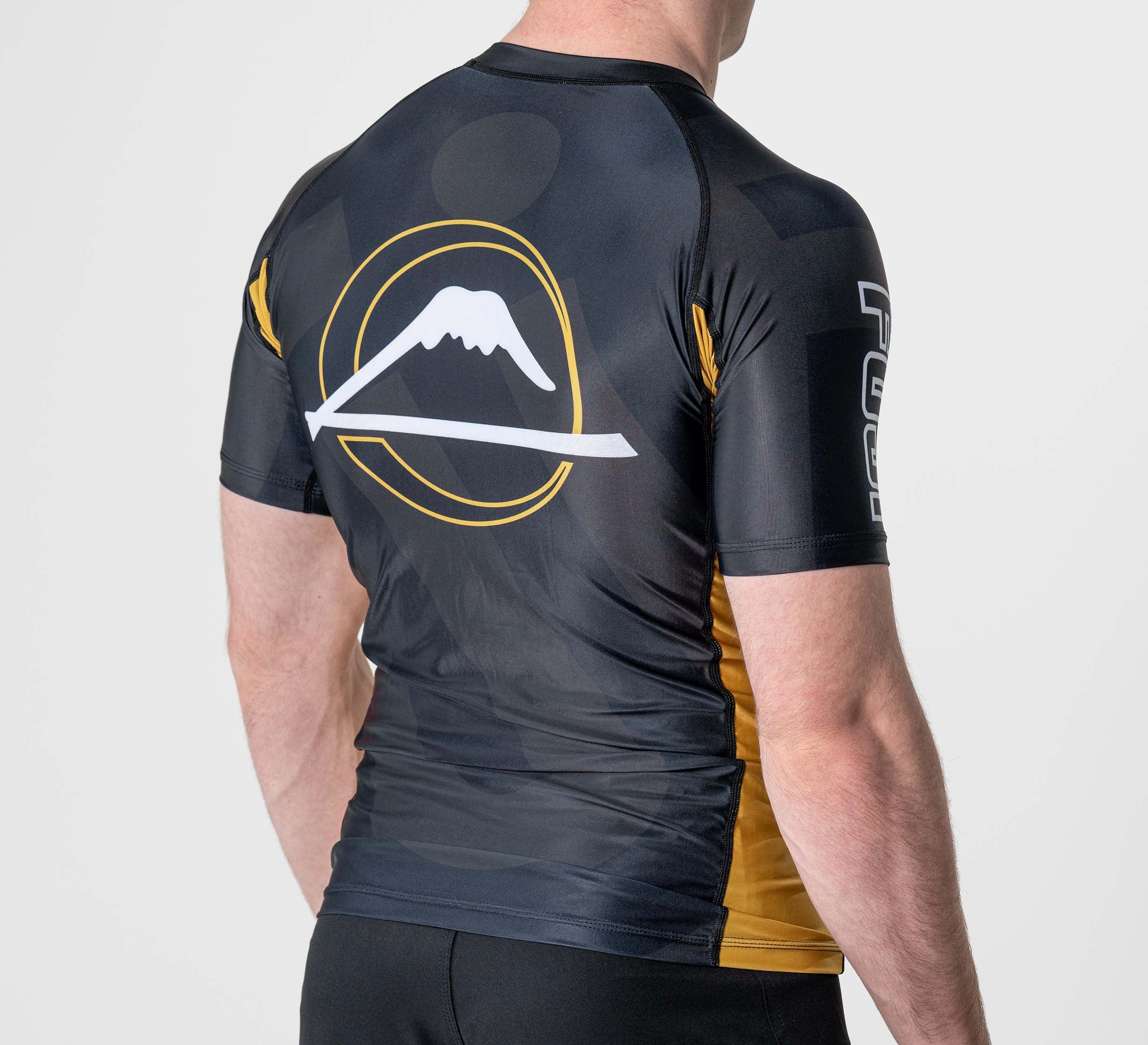 A person wearing the Rebel Flex Lite Rashguard Black, a black and yellow short-sleeve athletic shirt with a mountain and circle graphic on the back, stands against a plain light background.