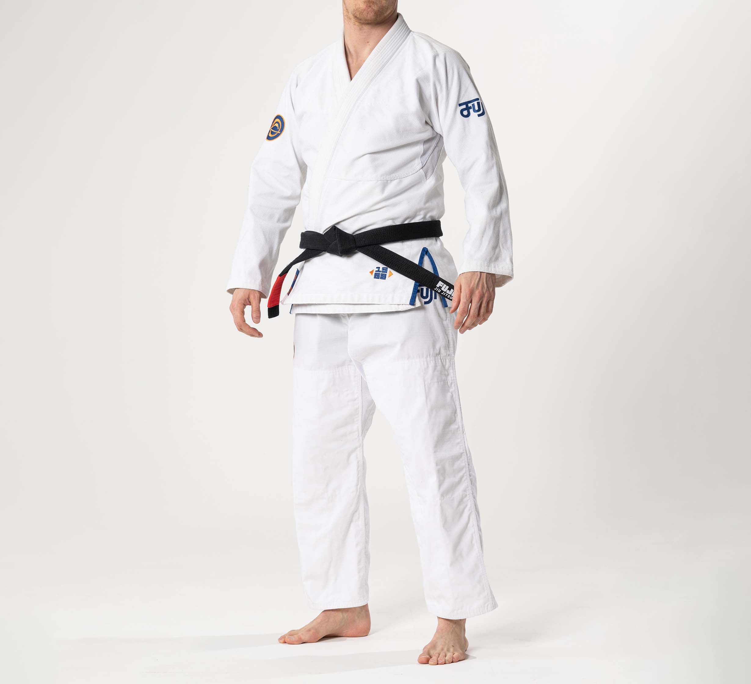 A person stands barefoot in a Flow-Tech BJJ Gi White/Navy/Orange with patches and a black belt, against a plain white background. Their face is not visible.