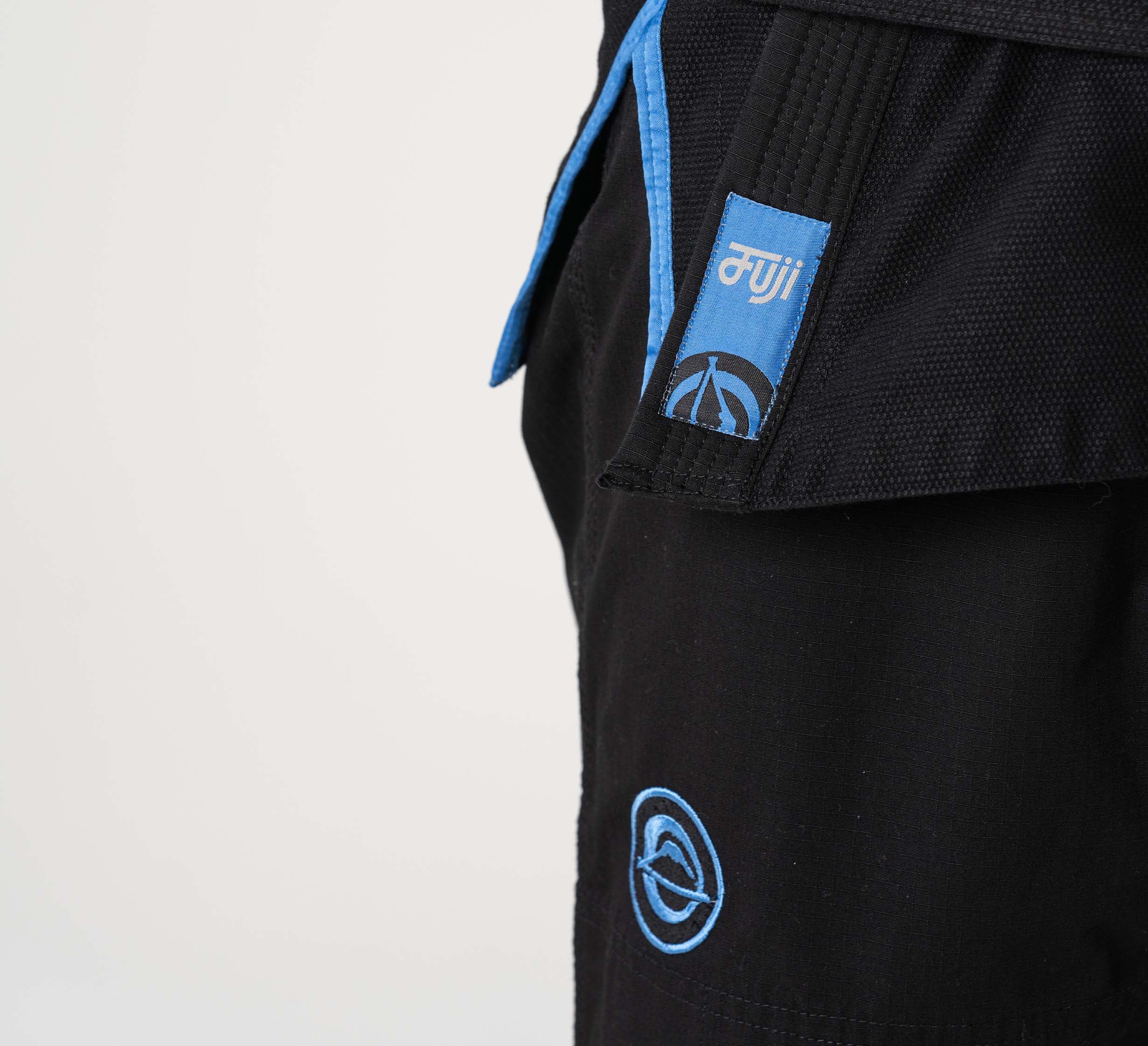 Close-up of the Flow-Tech BJJ Gi in black with blue trim, featuring embroidered logos: a white "muji" on a blue tab and a circular blue emblem on both the belt and pants.
