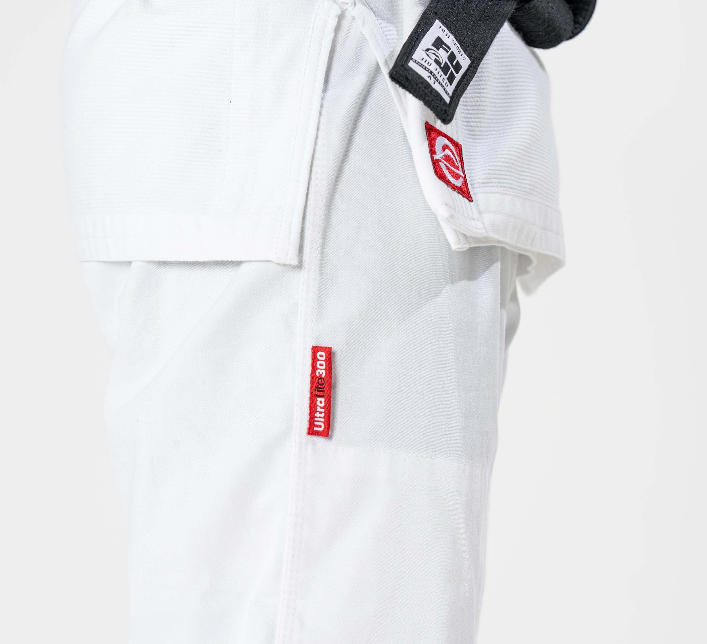 Close-up of a person wearing a white Ultra Lite 300 BJJ Gi with a black belt and a red tag on the pants; the gi also features an extra red patch on the jacket.