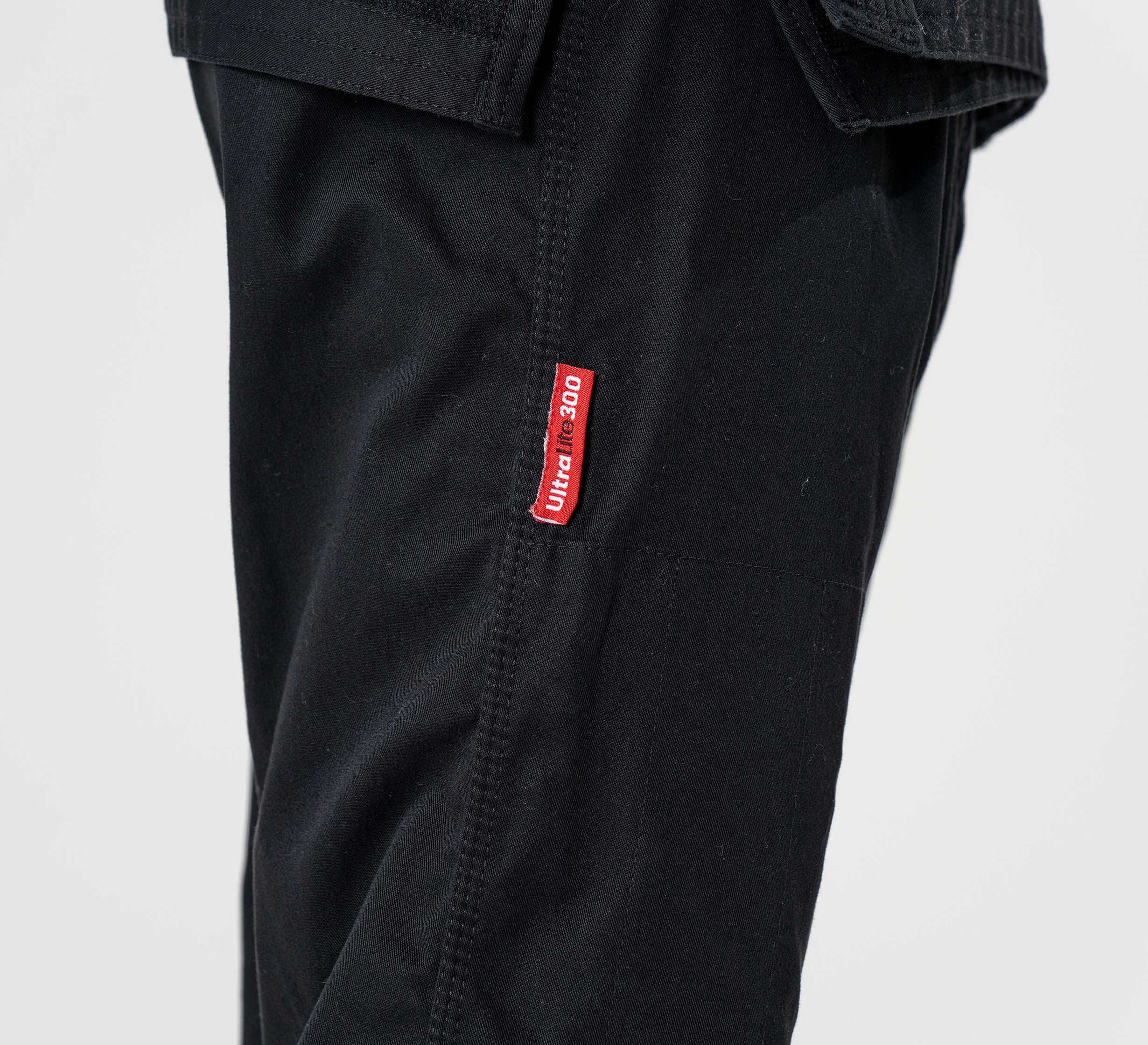 Close-up of a person wearing the Kids Ultra Lite 300 BJJ Gi Black, featuring a small red and white ULTIC 360 label sewn onto the left leg seam. The background is plain and light-colored.