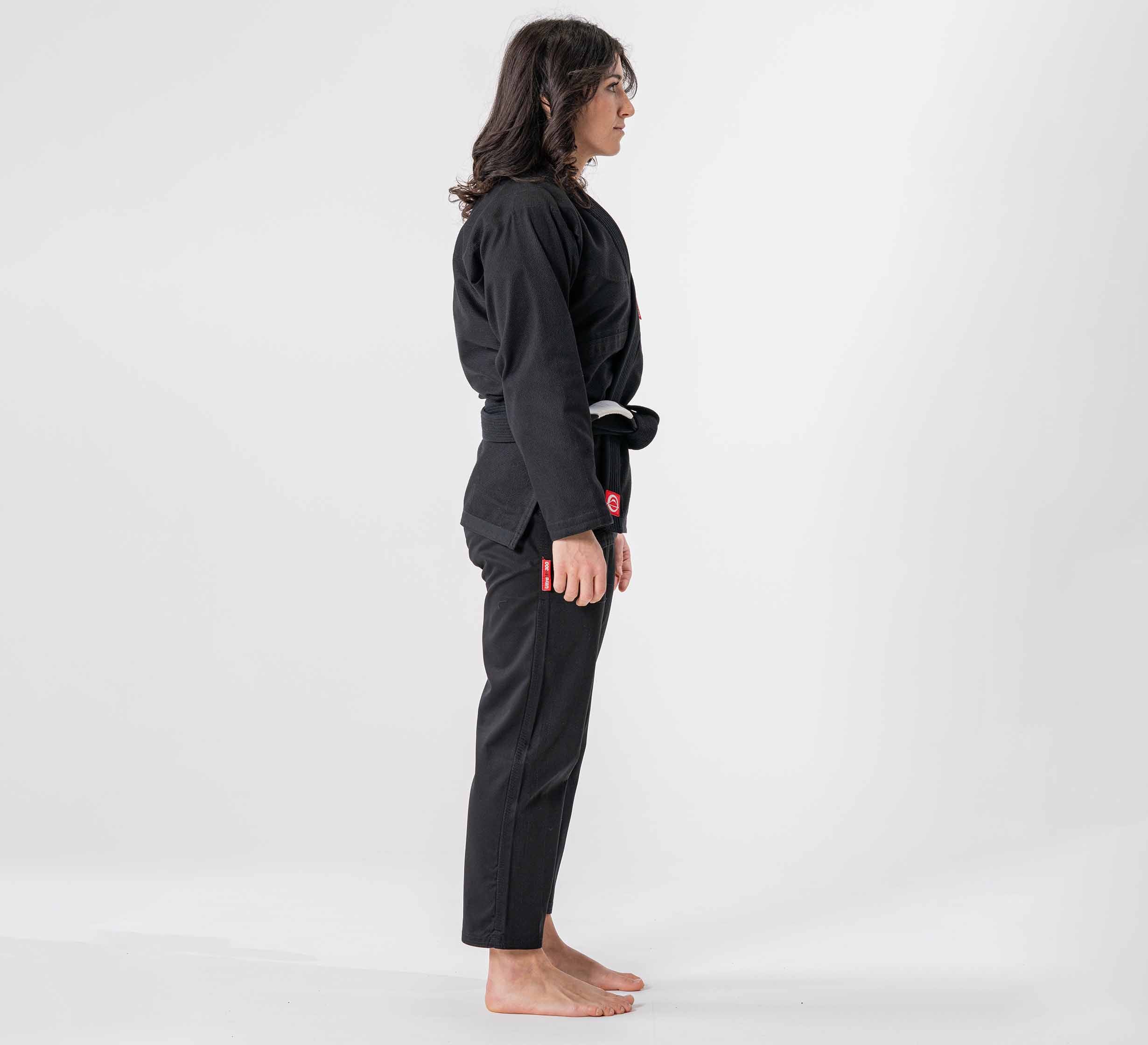 A woman with long dark hair stands barefoot in profile, wearing the Womens Ultra Lite 300 BJJ Gi Black with a black belt, against a plain white background.