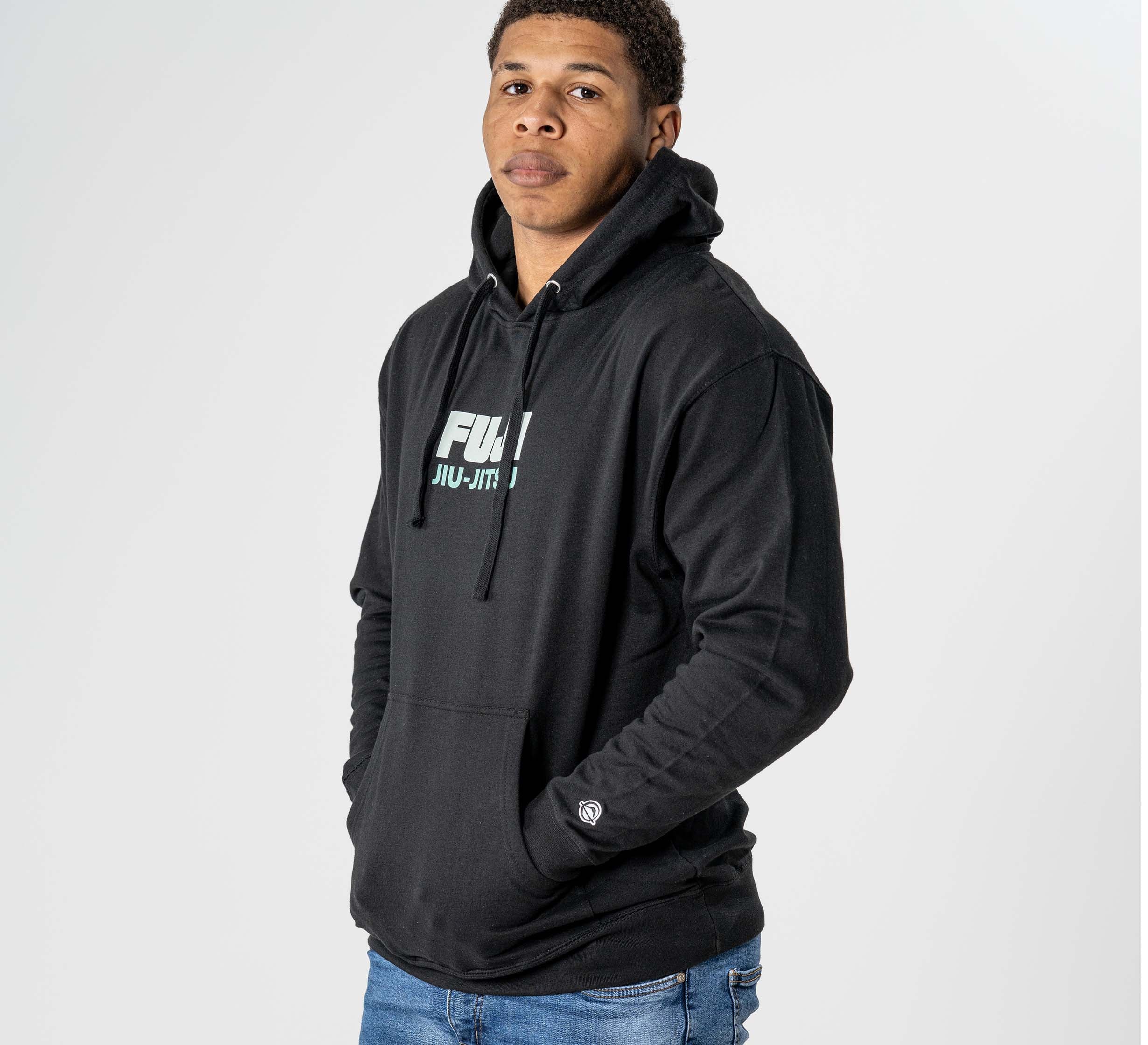 A man wearing the Black Belt Mentality Hoodie Black, featuring "FULL JIU-JITSU" on the front, stands against a plain white background with his hands in his pockets and blue jeans, looking at the camera.