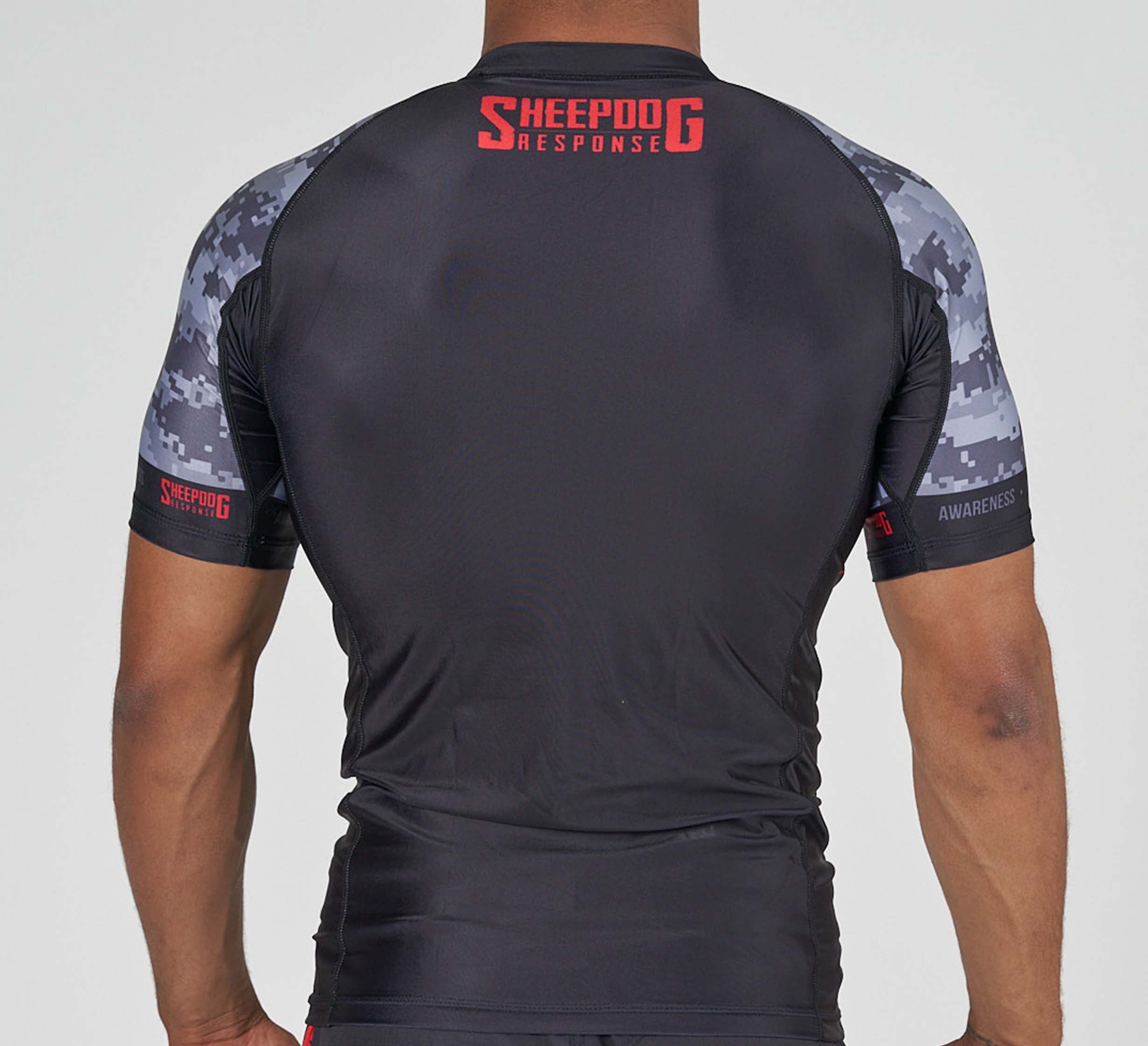 A person wears the Sheepdog Response Flex Lite Rashguard Black, featuring gray camouflage sleeves and "SHEEPDOG RESPONSE" in red letters near the neck on the back, with their arms at their sides.