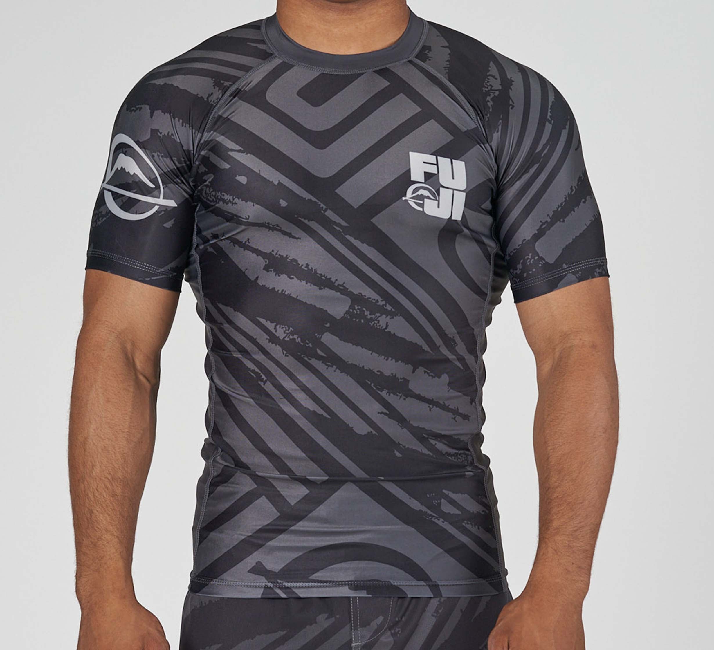 A person wears the Battle Flex Lite Rashguard Black, a fitted, short-sleeve athletic top with black and gray patterns and “FUJI” with a mountain logo on the chest and sleeve, standing against a plain light background.