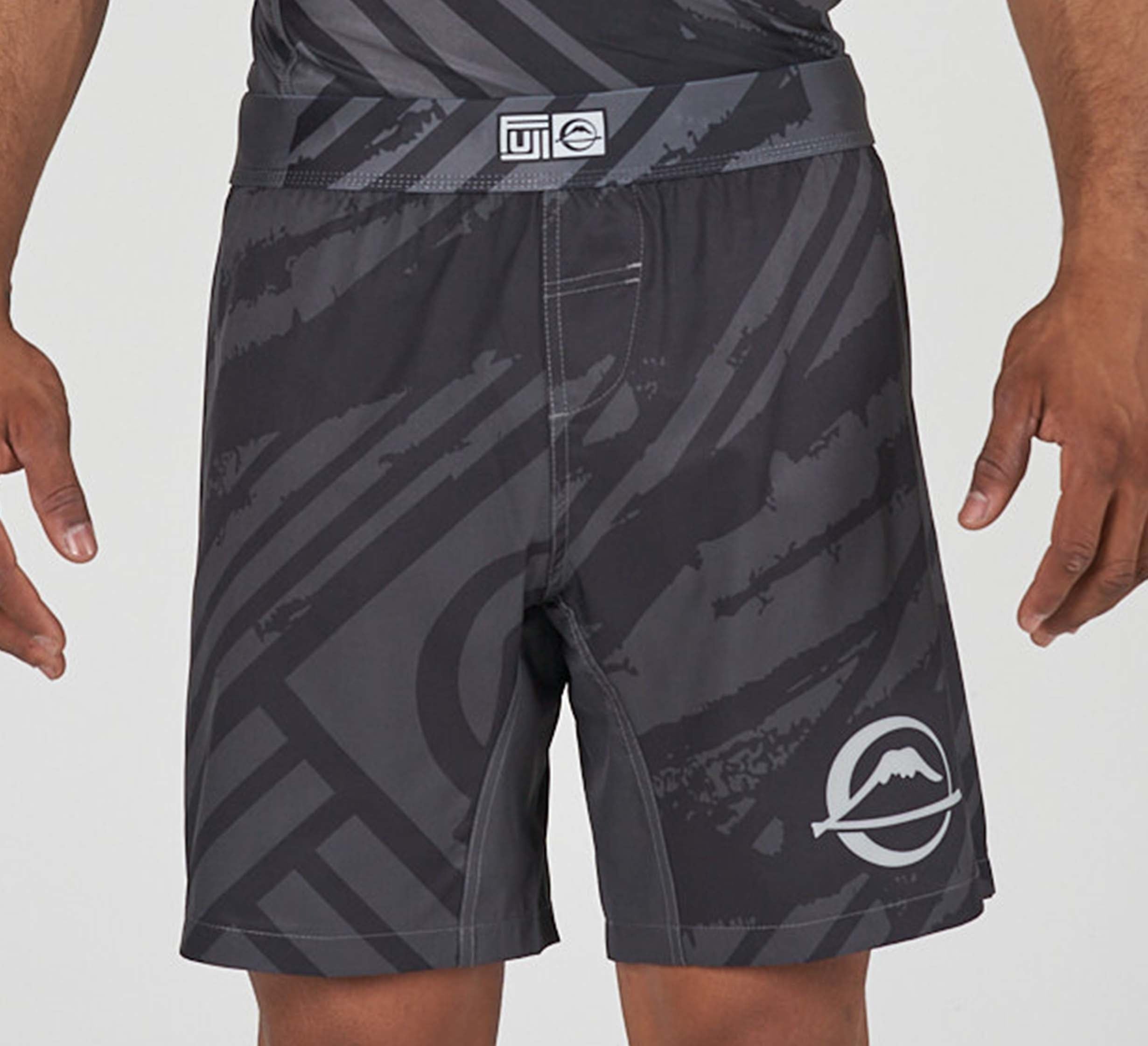 A person wearing Battle Flex Lite Shorts Black, featuring black and gray abstract patterns and a mountain logo on the left leg. Both hands are visible at their sides.