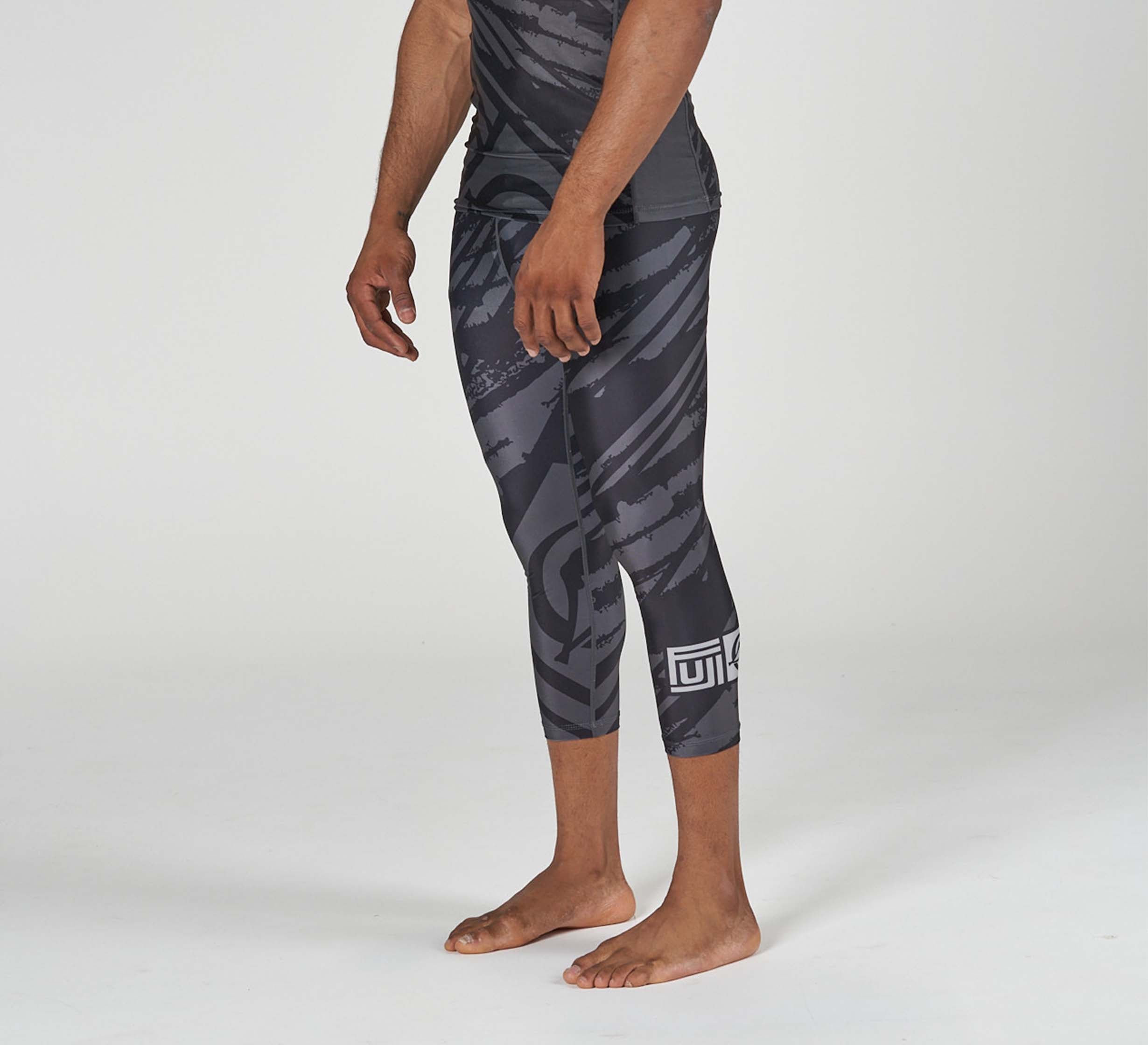 A person stands barefoot wearing Battle Flex Lite Spat Black compression leggings with black and gray patterns and white geometric designs on the lower leg, against a plain white background.
