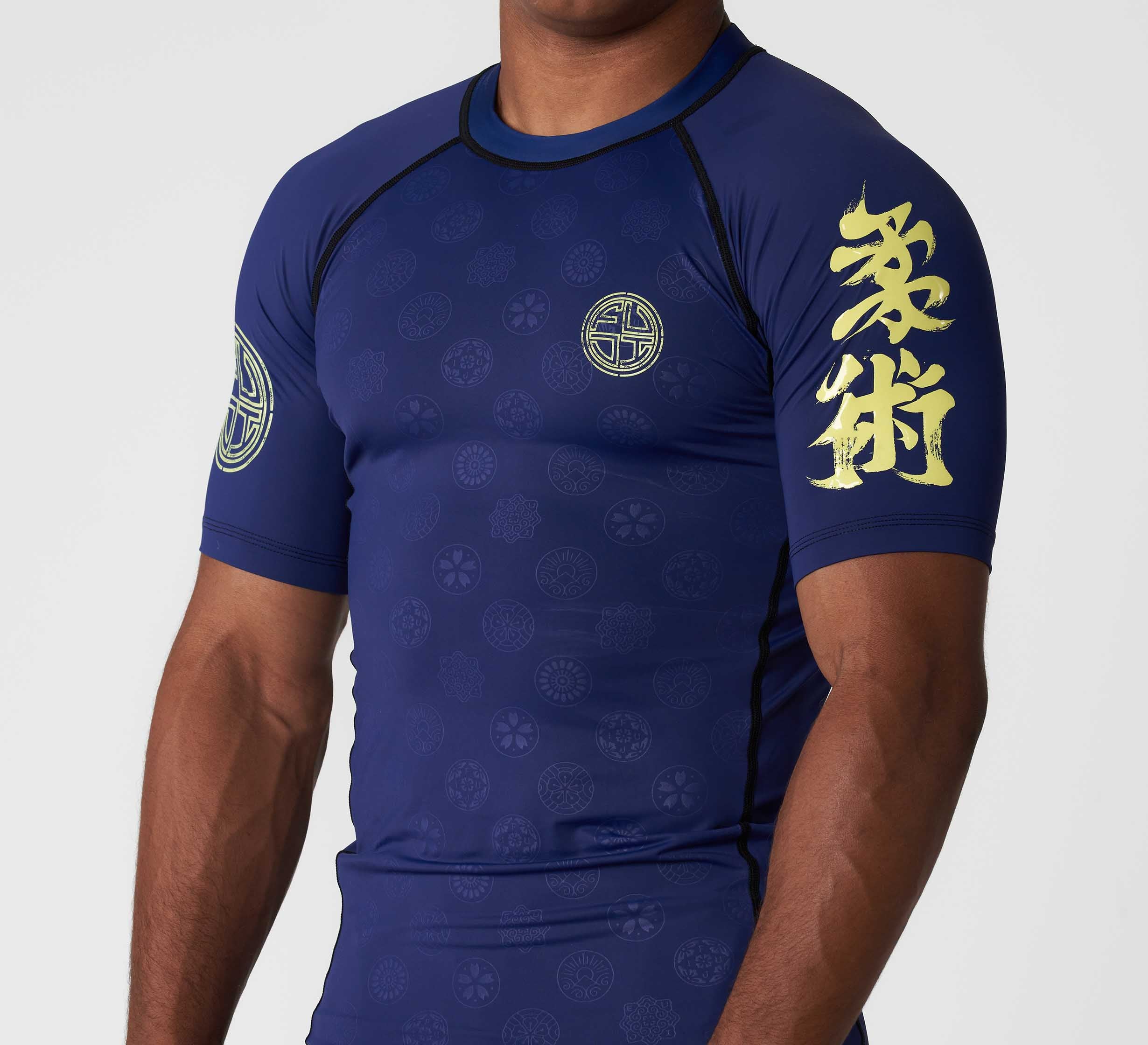 A man wears the Shogun Heat Gear Rashguard Navy, a short-sleeved navy top with gold Japanese kanji on the left sleeve and circular logos on the front and right sleeve.