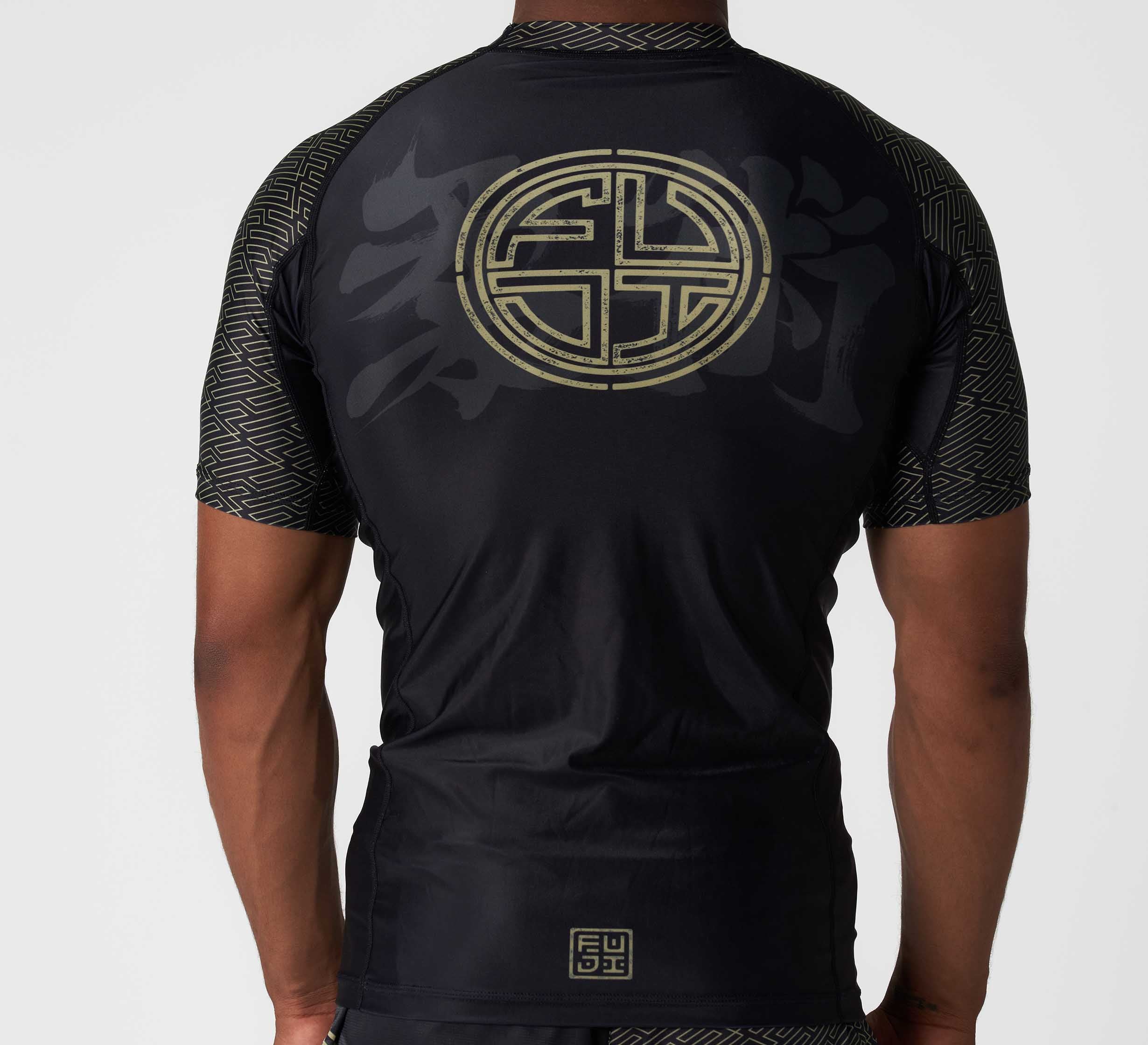 A person shown from behind wears the Bushido Flex Lite Rashguard, a black short-sleeve athletic shirt featuring geometric patterns and a large circular gold Asian-inspired design on the back, set against a plain background.