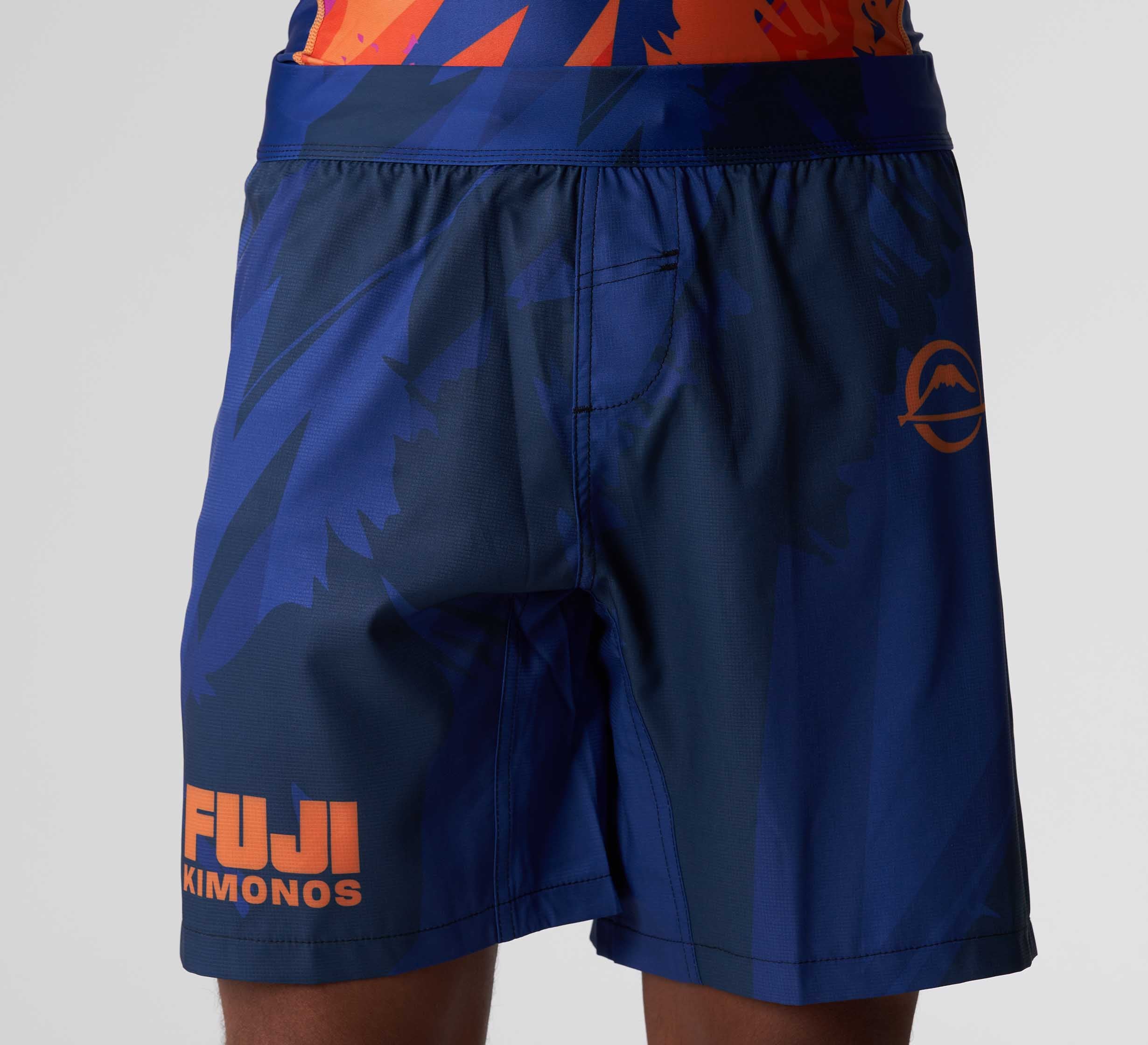 A person wearing Hanzo Flex Lite Shorts Blue featuring an abstract pattern, orange accents, "FUJI KIMONOS" text, and a small logo on the left leg.