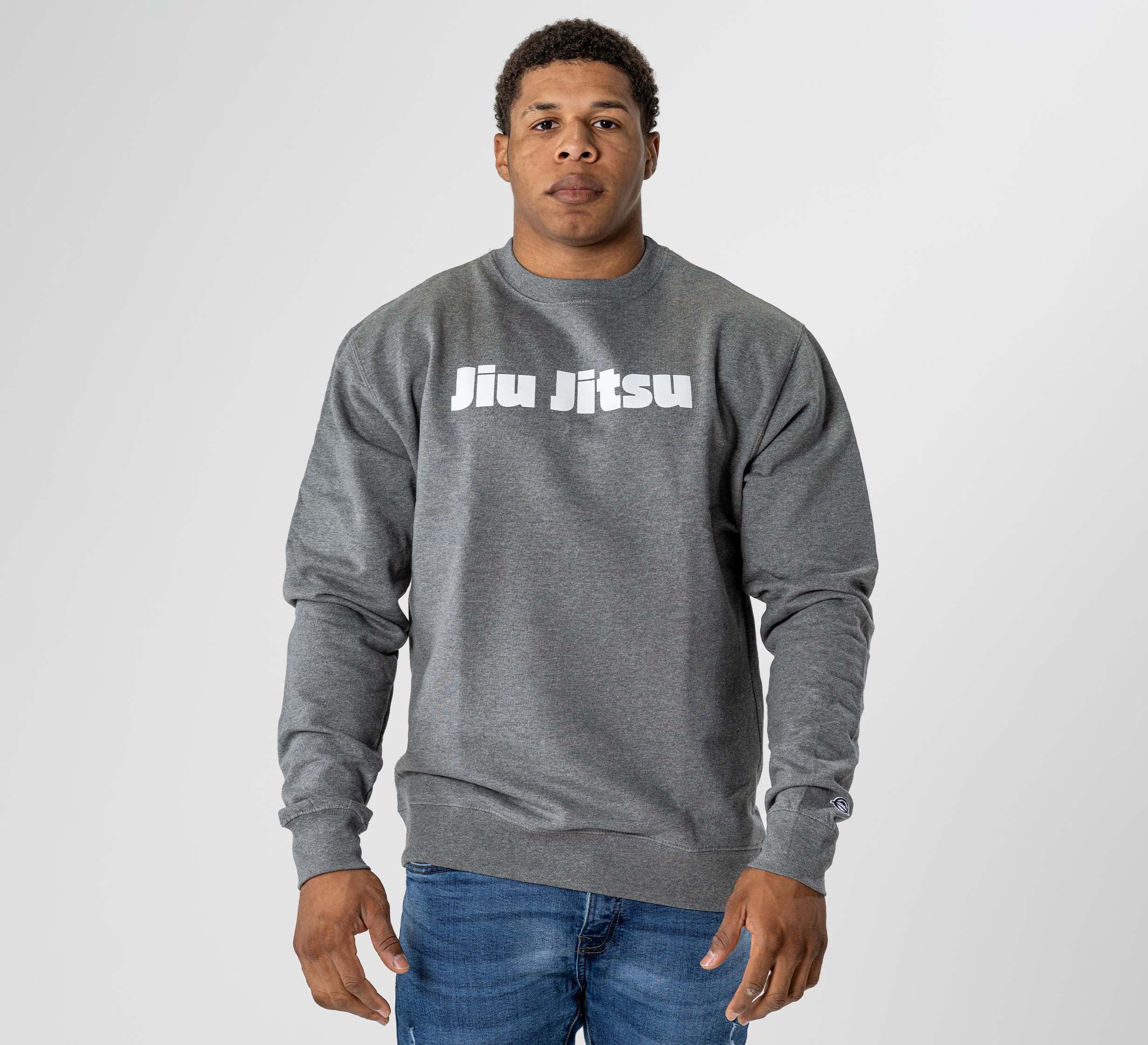 A man faces forward in a Jiu Jitsu Player Crewneck Heather Grey sweatshirt with bold white "Jiu Jitsu" lettering, paired with blue jeans, set against a plain light gray background.