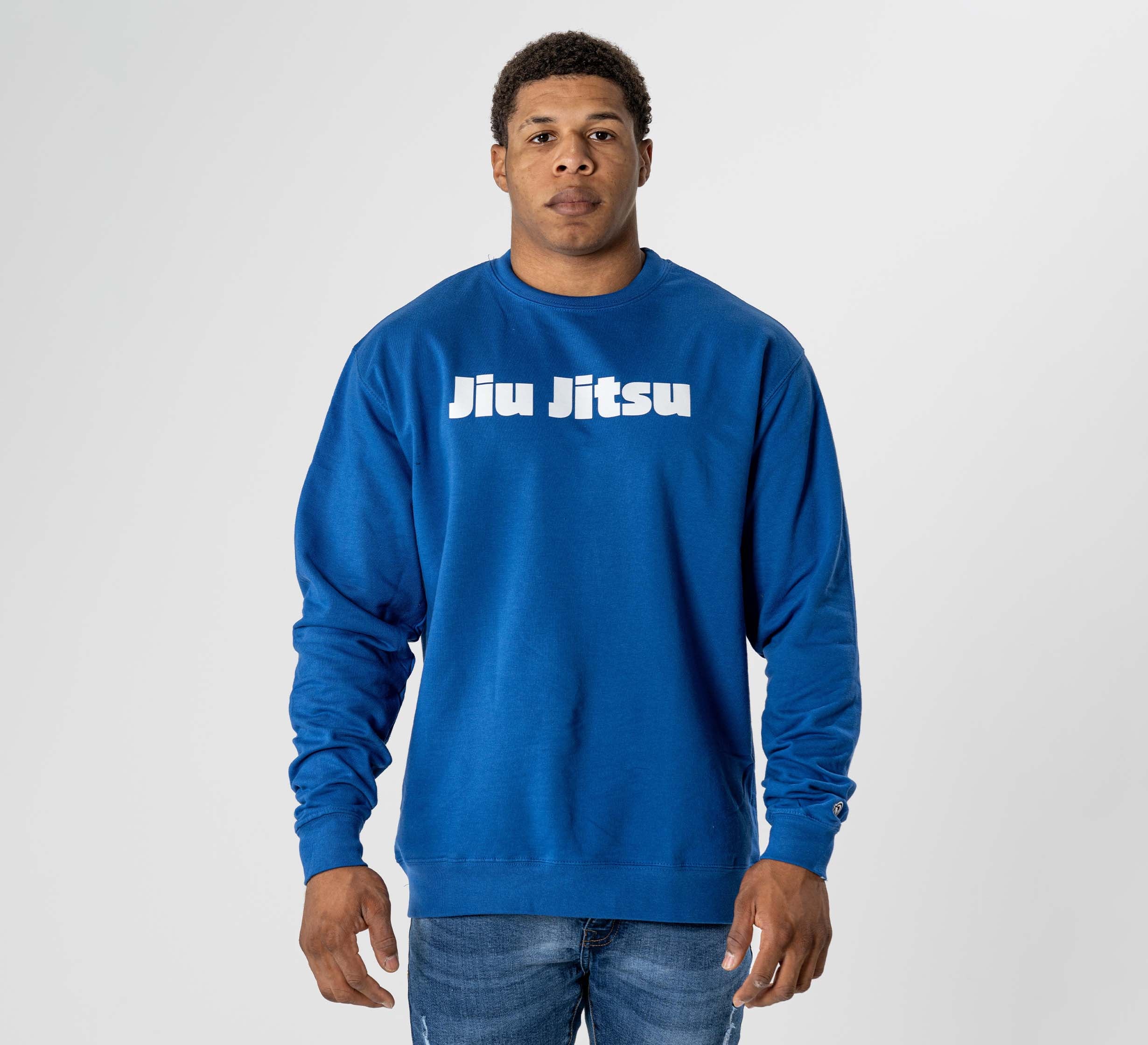 A man faces forward in the Jiu Jitsu Player Crewneck Blue sweatshirt with "Jiu Jitsu" in bold white letters on the front, paired with blue jeans, against a plain light gray background.