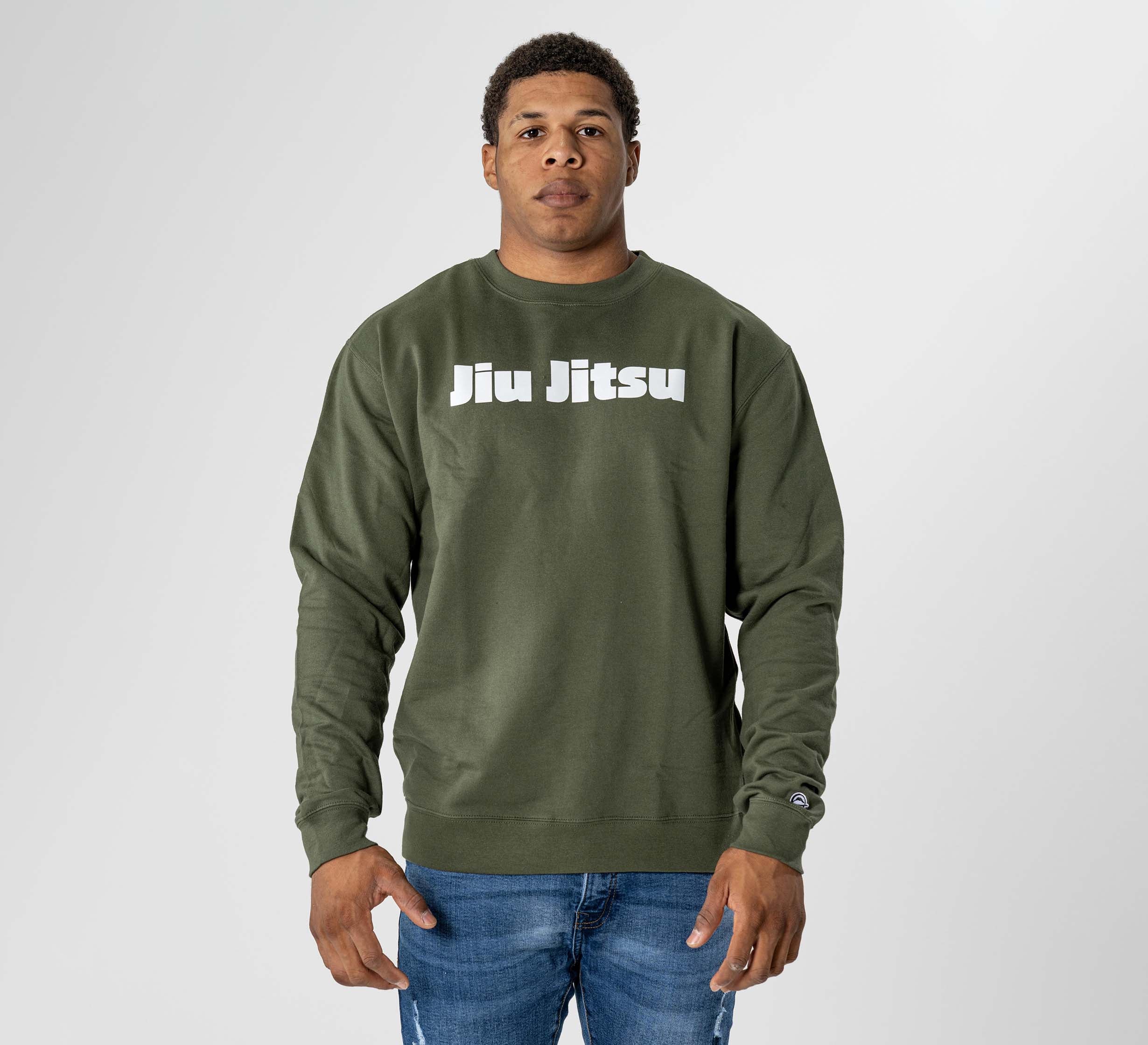 A person stands facing forward in the Jiu Jitsu Player Crewneck Military Green, featuring bold white "Jiu Jitsu" lettering, paired with blue jeans against a plain light background.