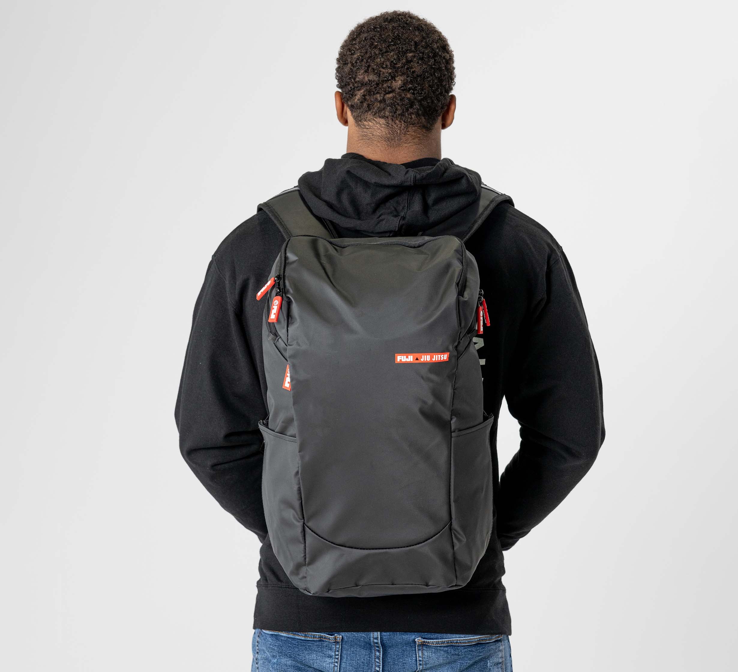 A person in a black hoodie and blue jeans is seen from behind carrying the Urban Day Backpack Black, featuring red zipper pulls and a small red and white logo on the back.