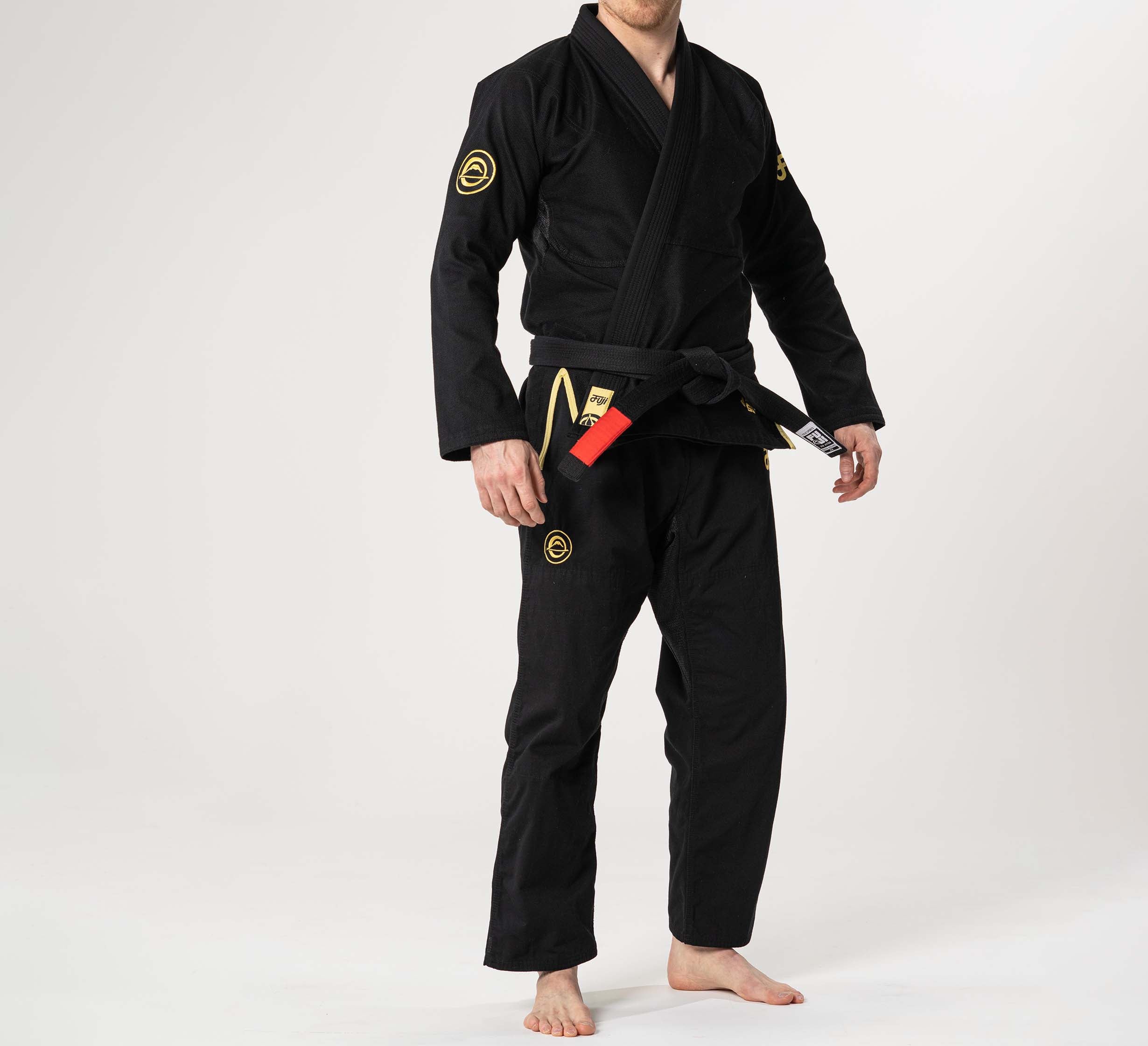 A person wearing the Flow-Tech BJJ Gi Black/Gold and a black belt stands barefoot against a plain white background, with their face partially out of frame.