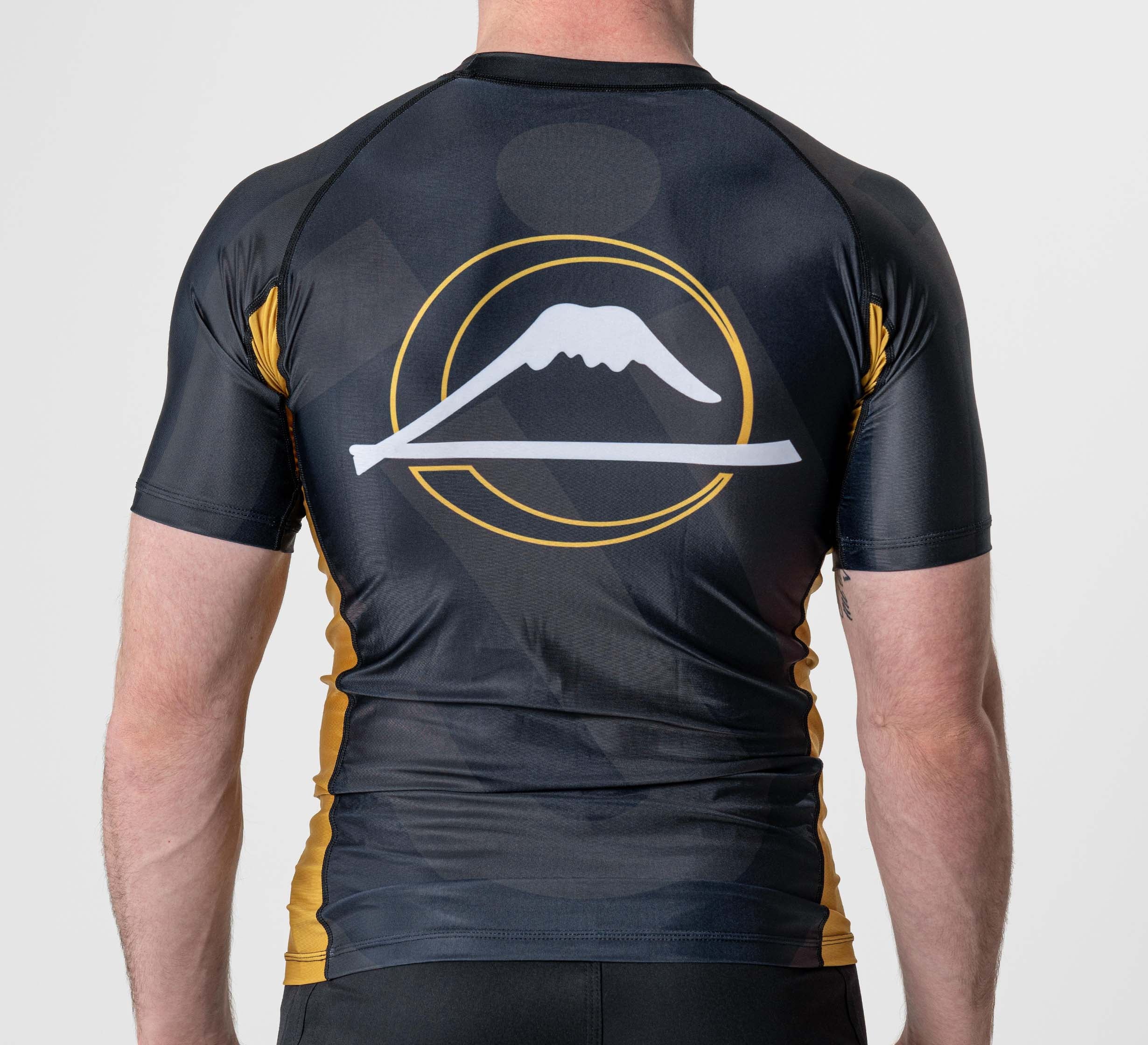 A person is seen from behind wearing the Rebel Flex Lite Rashguard Black, a fitted short-sleeve athletic shirt with a white and yellow mountain graphic above a line inside a circle on the back.